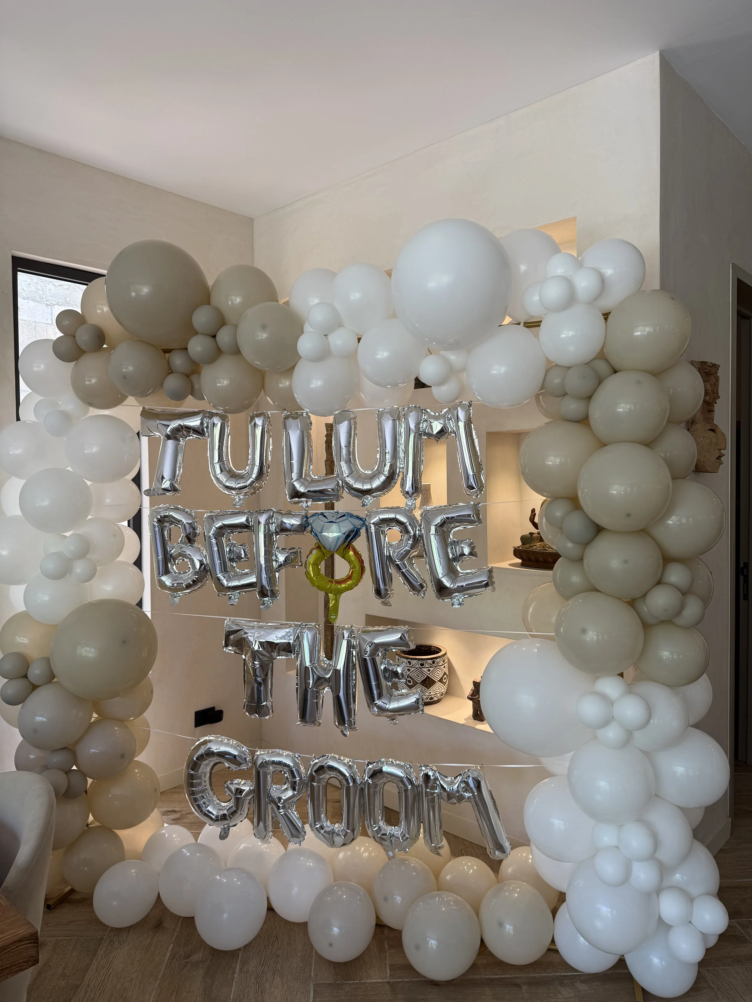 Decorative arch made for a Bachelorette of white and beige balloons with silver balloons spelling out 'TULUM BEFORE THE GROOM' and additional foil balloons.
