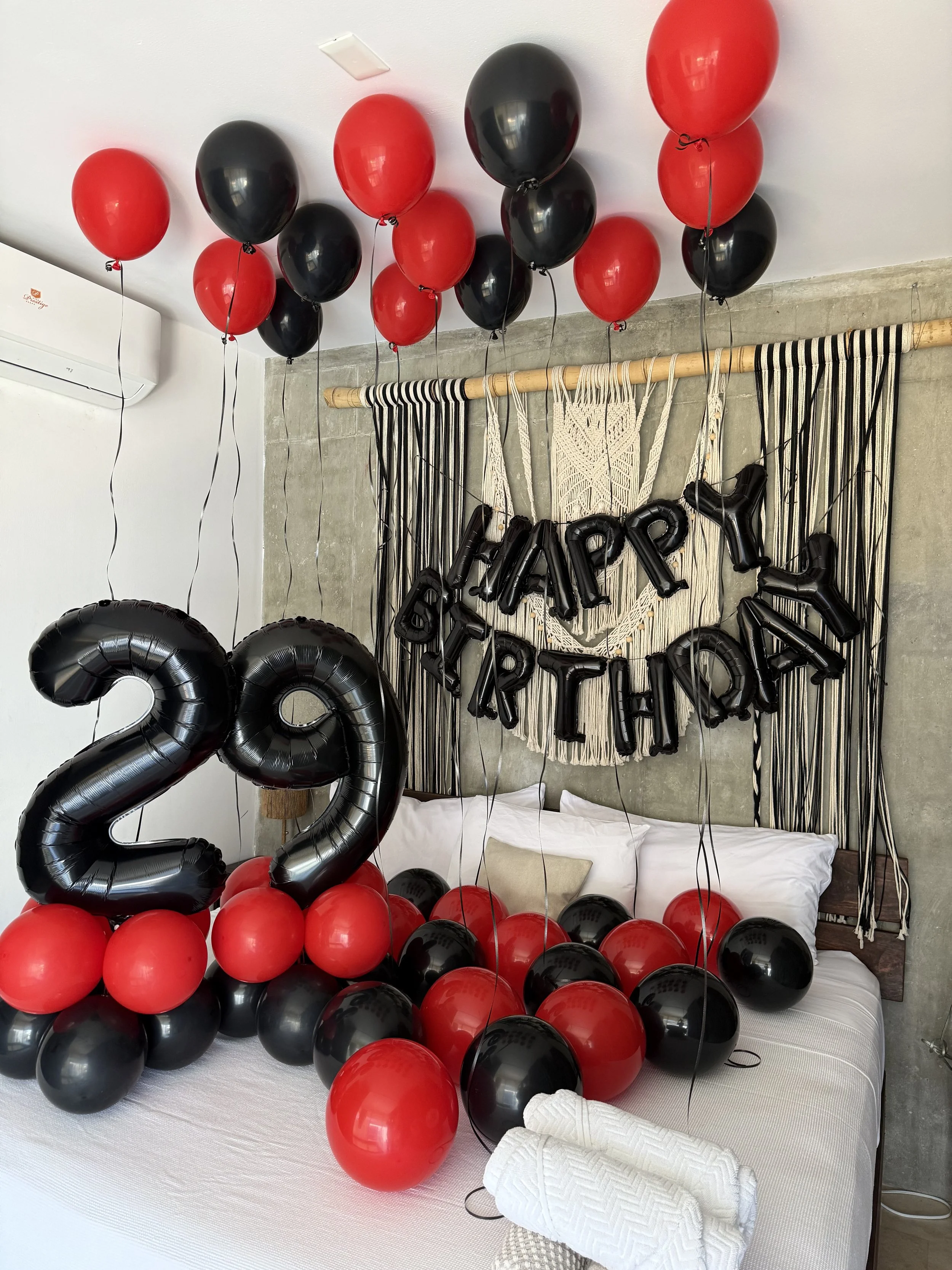 Decorations for a 2022 celebration including black and red balloons, some in the shape of the year 2022, arranged on a bed with white sheets, pillows, and towels, with a macramé wall hanging in the background.
