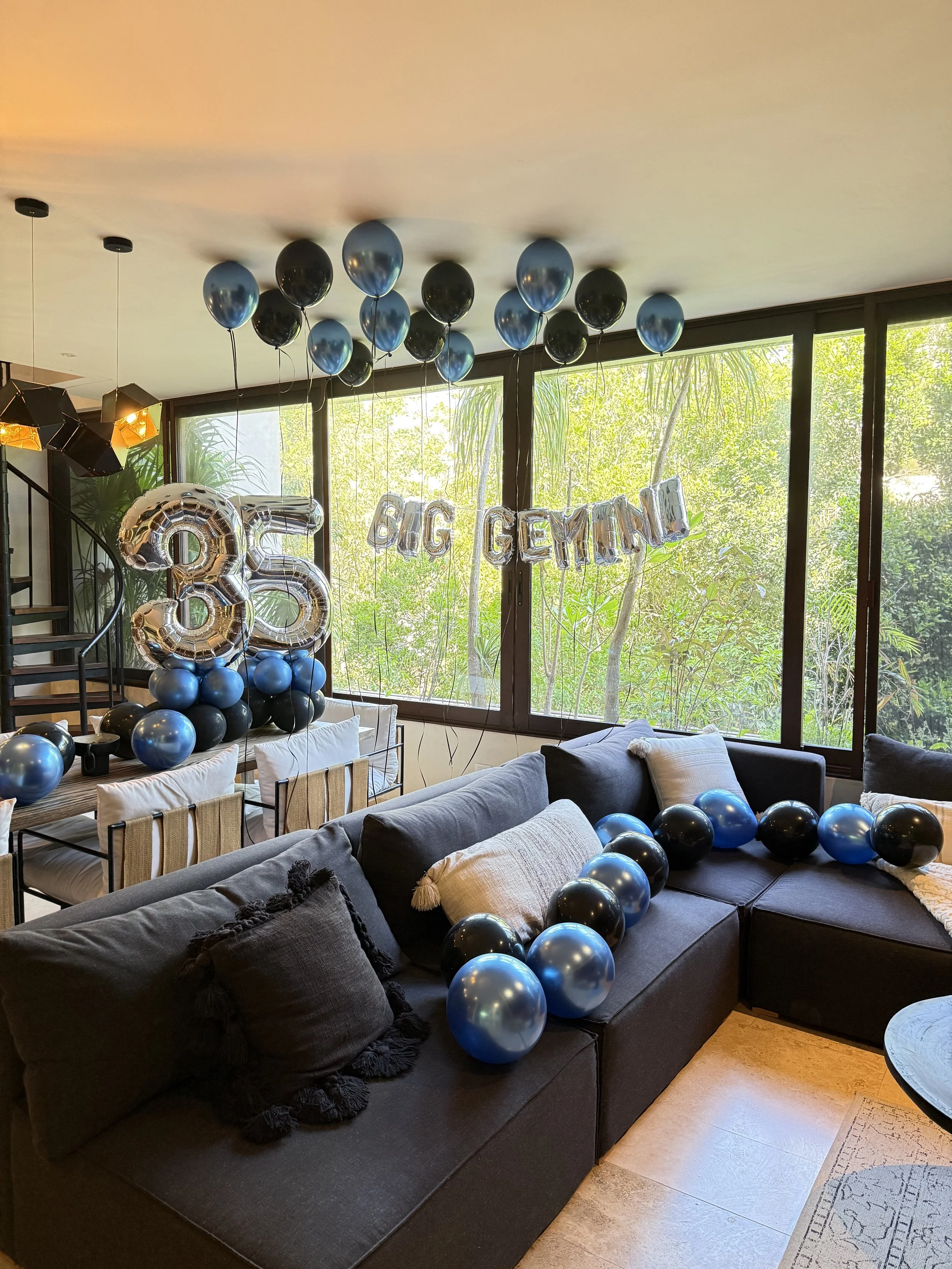 Living room decorated with black and blue balloons, including large number balloons '3' and '5', and silver 'BIG GERMAN' balloons, for a 35th birthday celebration, with large windows and green trees visible outside.