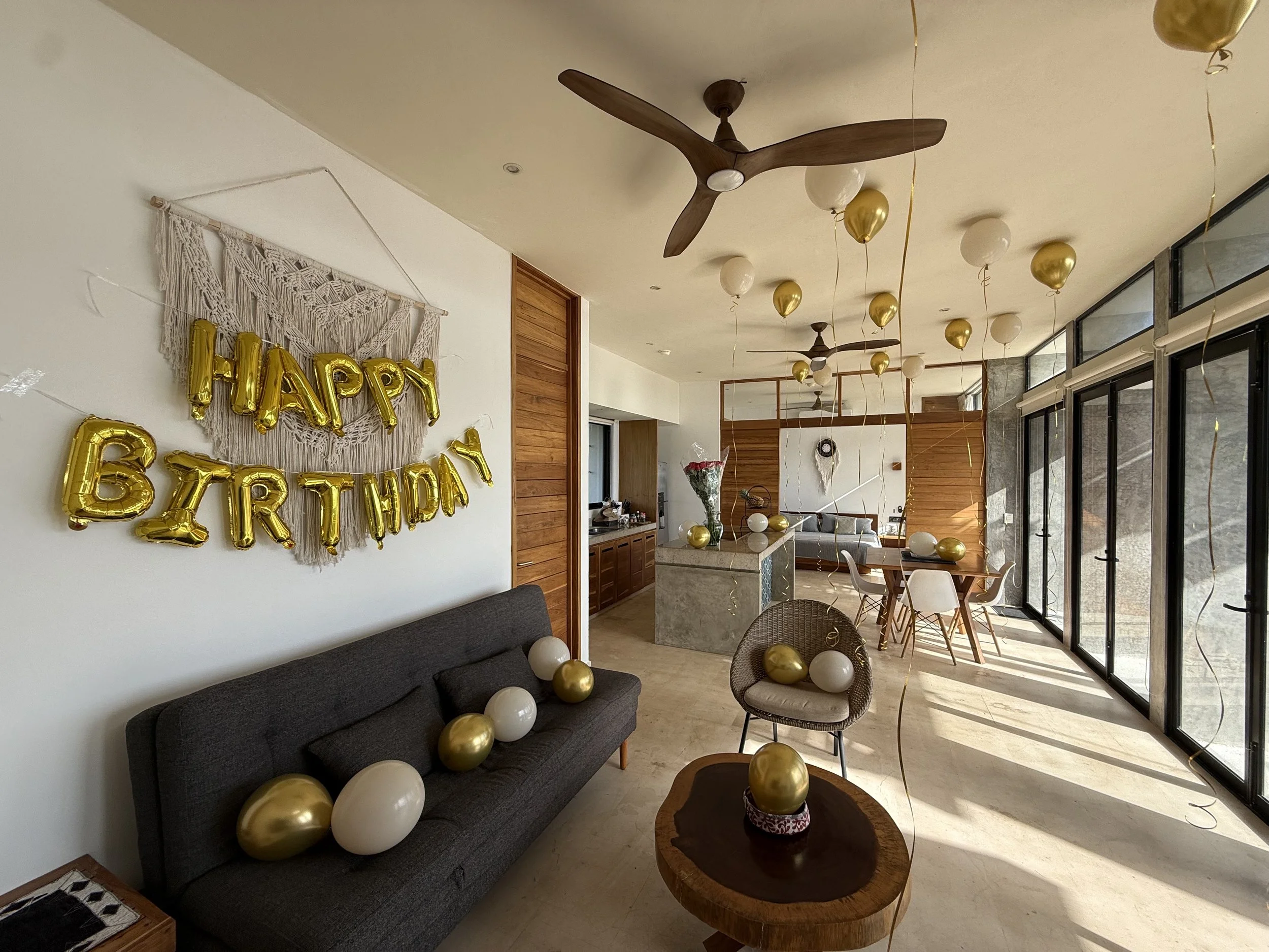 Living room decorated with gold and white balloons for a birthday celebration, including a "Happy Birthday" banner on the wall, a gray sofa, and large glass windows letting in natural light.