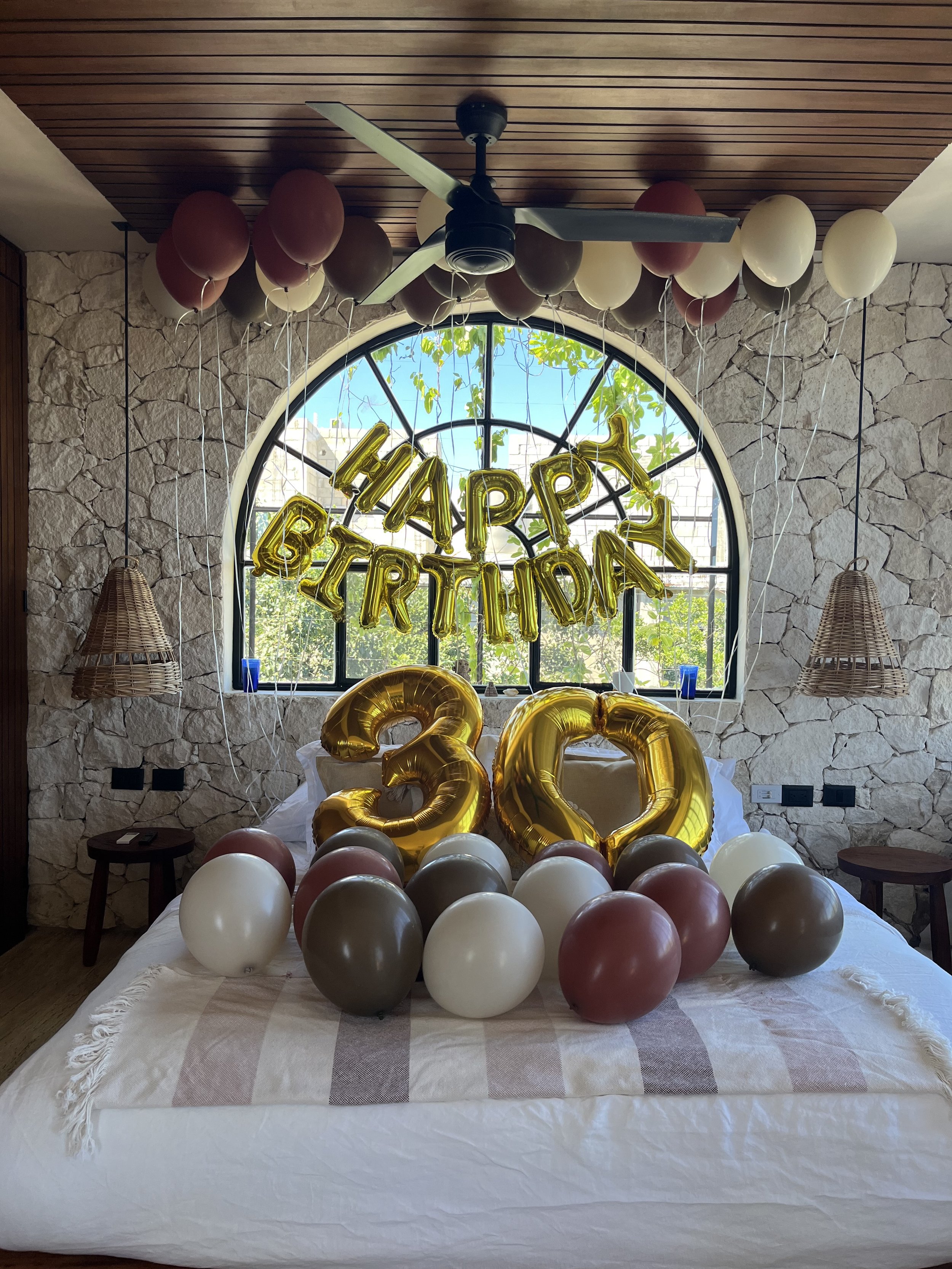Decorated bedroom with balloons for a 30th birthday celebration, featuring a 'Happy Birthday' gold balloon banner, large gold number balloons '30', and surrounding balloons in white, brown, and maroon.