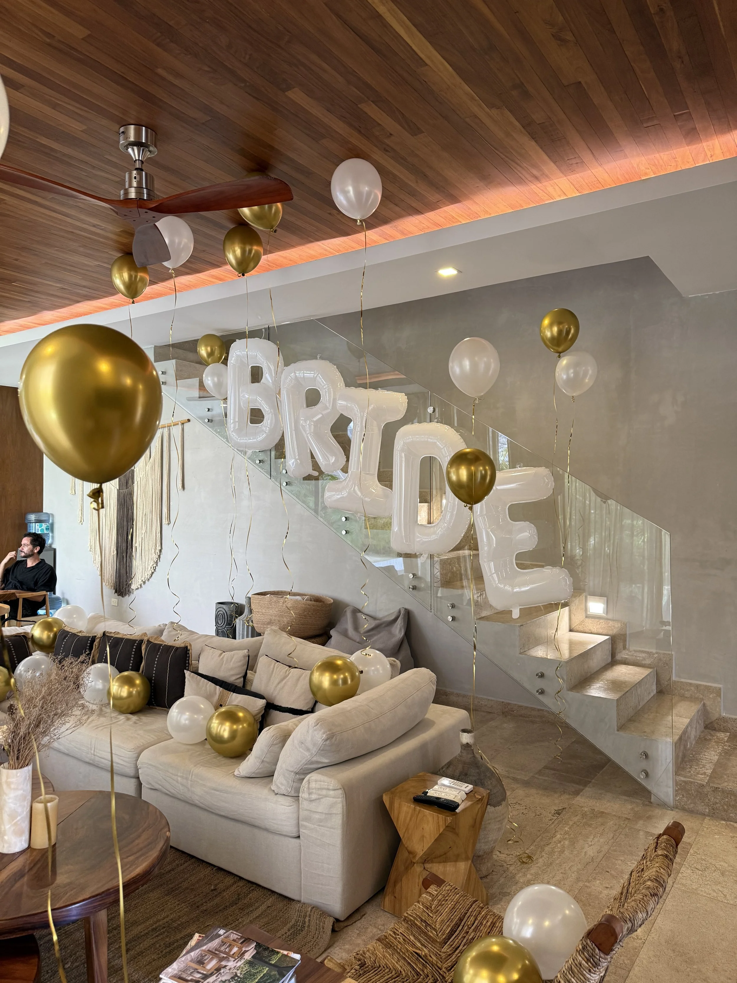 Living room decorated with white and gold balloons, large white balloons spelling out 'BRIDE' hanging from the ceiling, a beige sectional sofa with pillows, a wooden side table with some remote controls, a round wooden coffee table with balloons on it, a staircase with glass railing behind, Caucasian man sitting on a chair in the background, and balloons scattered across the room.
