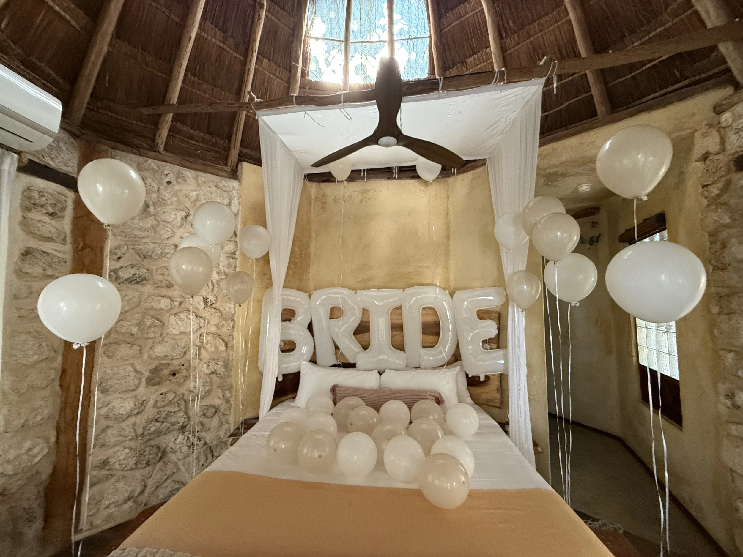White balloons on a bed with a 'BRIDE' balloon sign on the headboard, decorated for a bridal celebration or bachelorette party.