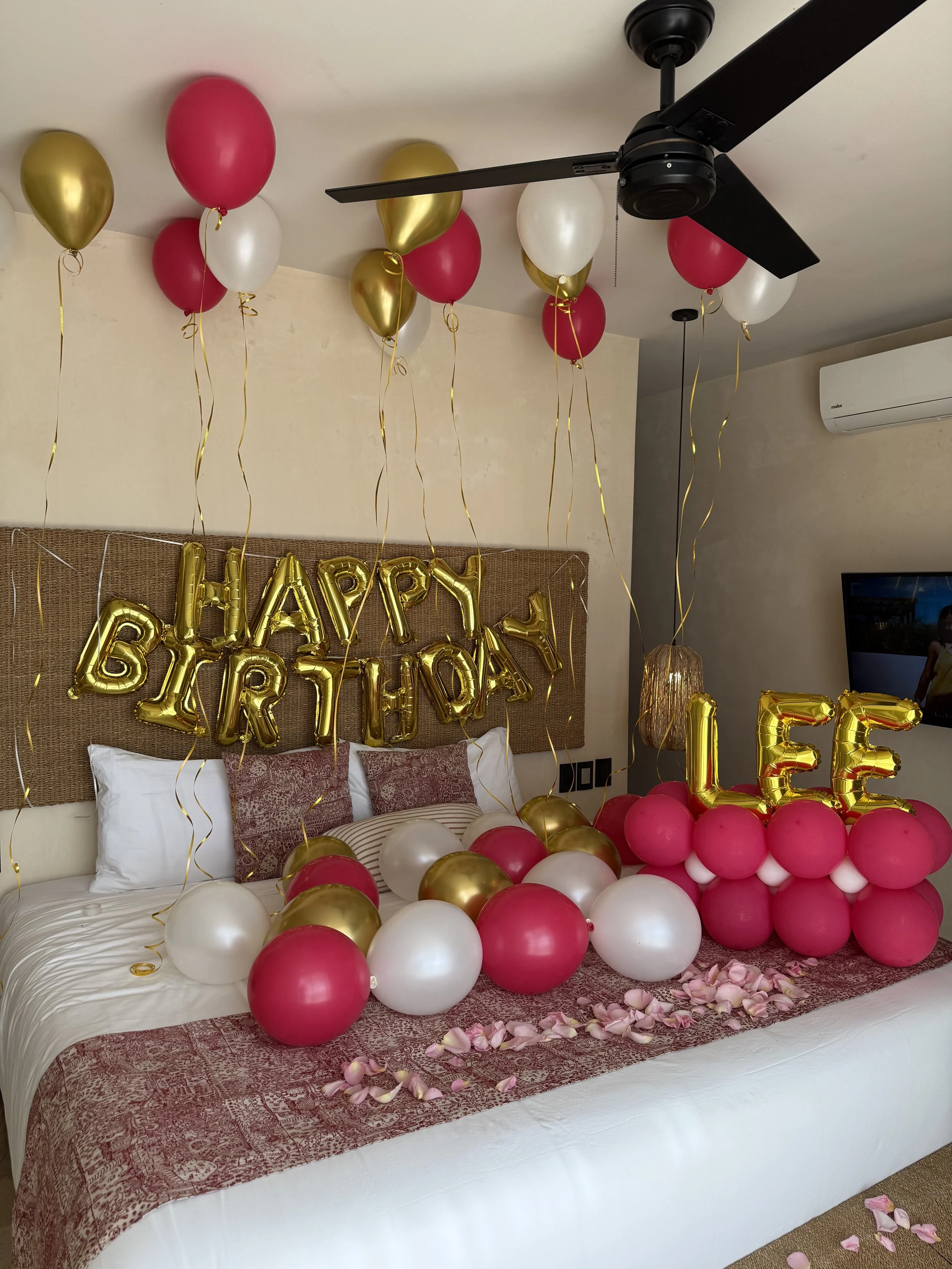 Decorated hotel room for a birthday celebration with pink, white, and gold balloons, a 'Happy Birthday' banner, and balloons spelling 'LEE' on the bed, with rose petals scattered.