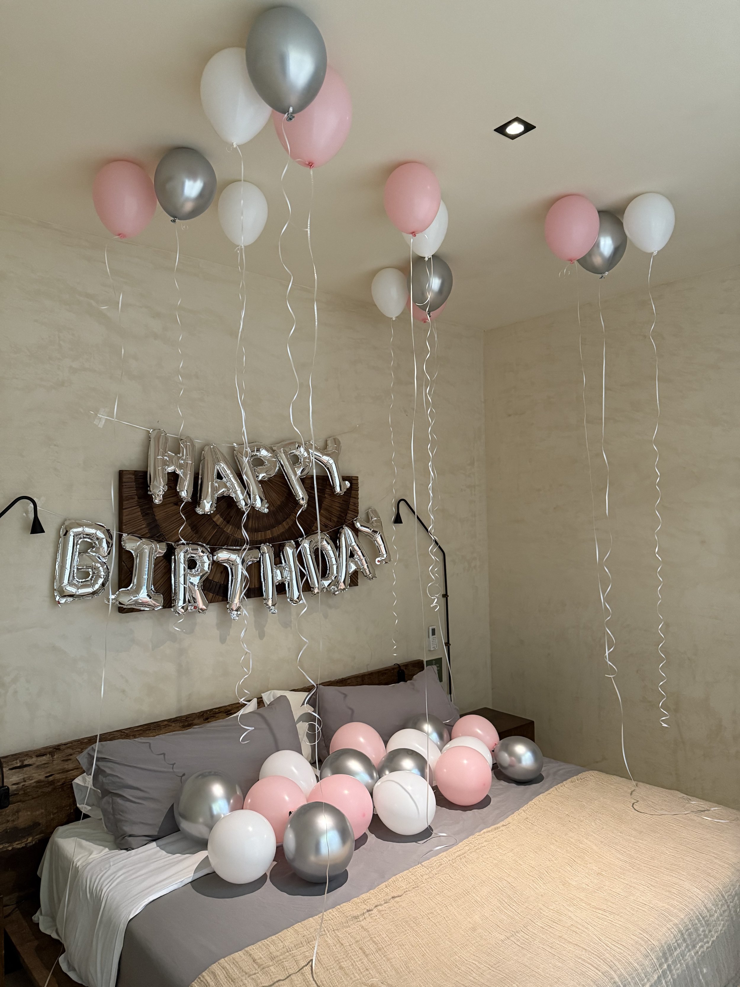 Decorated bedroom with pink, white, and silver balloons, a 'Happy Birthday' balloon banner on the wall, and a bed with pillows and balloons scattered on it.