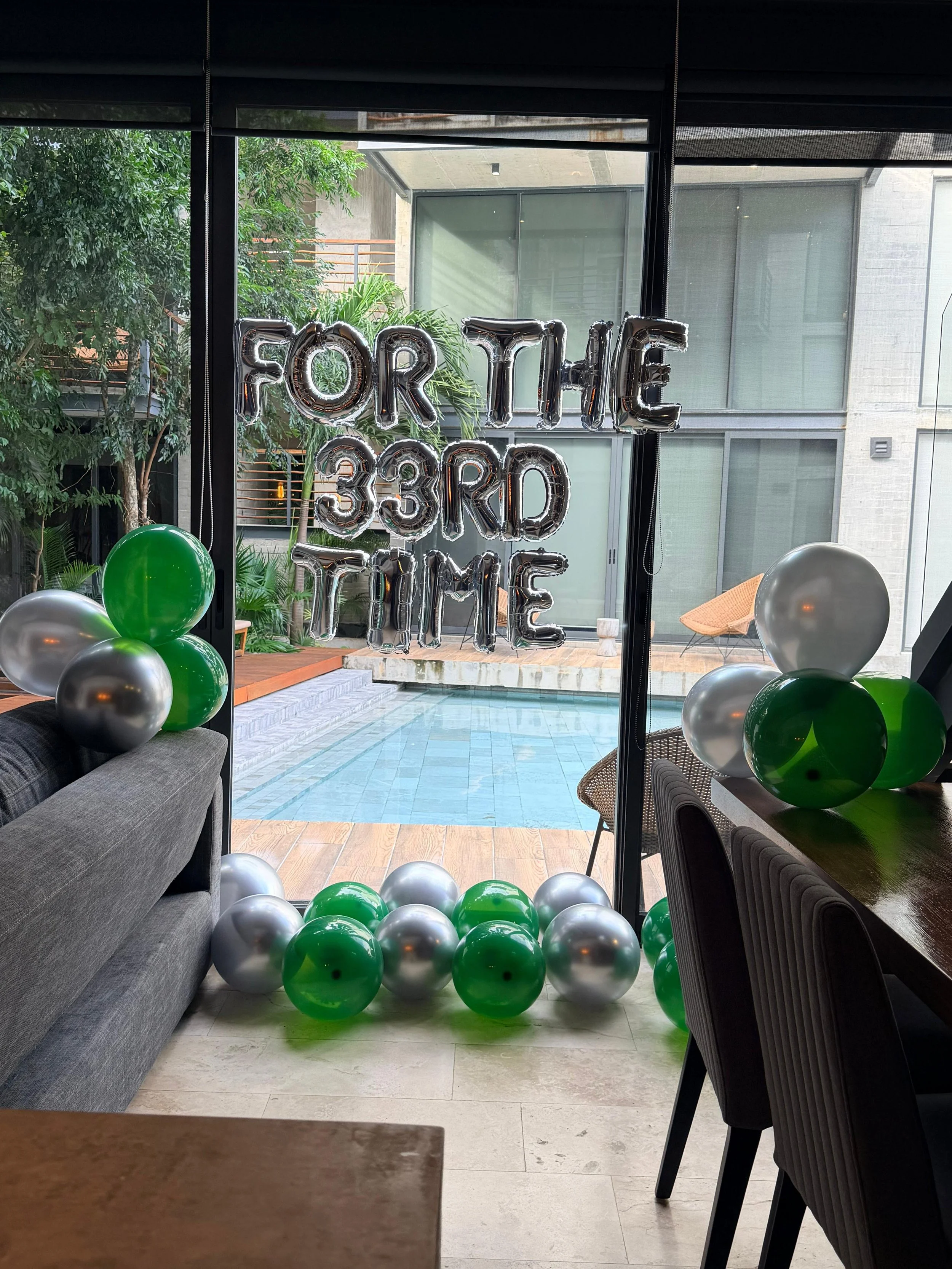 Indoor celebration with silver, green, and gray balloons, and a silver balloon sign that reads 'FOR THE 33RD TIME.' The view looks out to a patio with a pool and modern building exterior in the background.