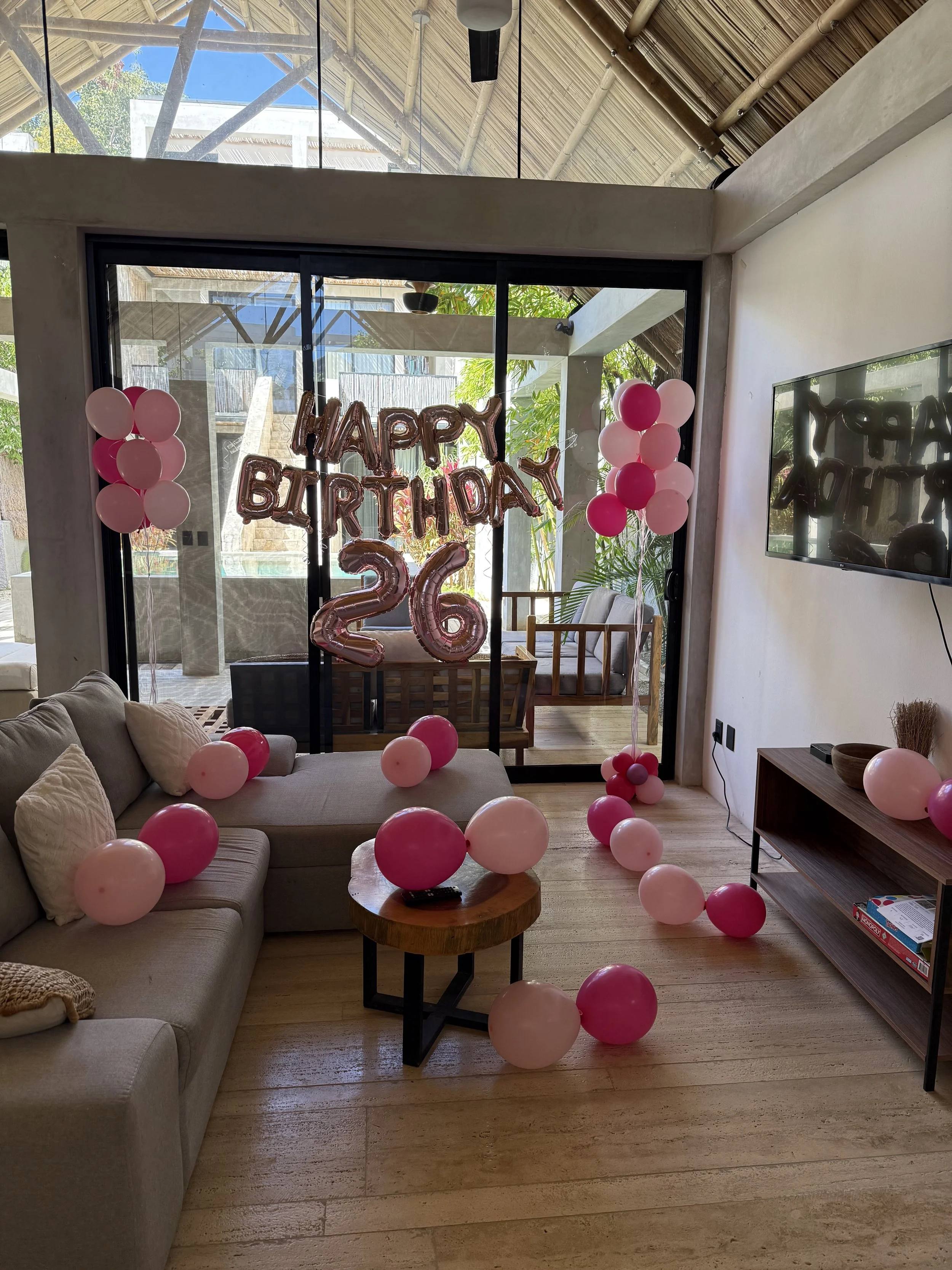 Living room decorated for a birthday celebration with pink and white balloons, a 'Happy Birthday' banner, and large number '26' balloons. There is a couch, a small round table, a TV on the wall, and a glass door leading to an outdoor area with wooden