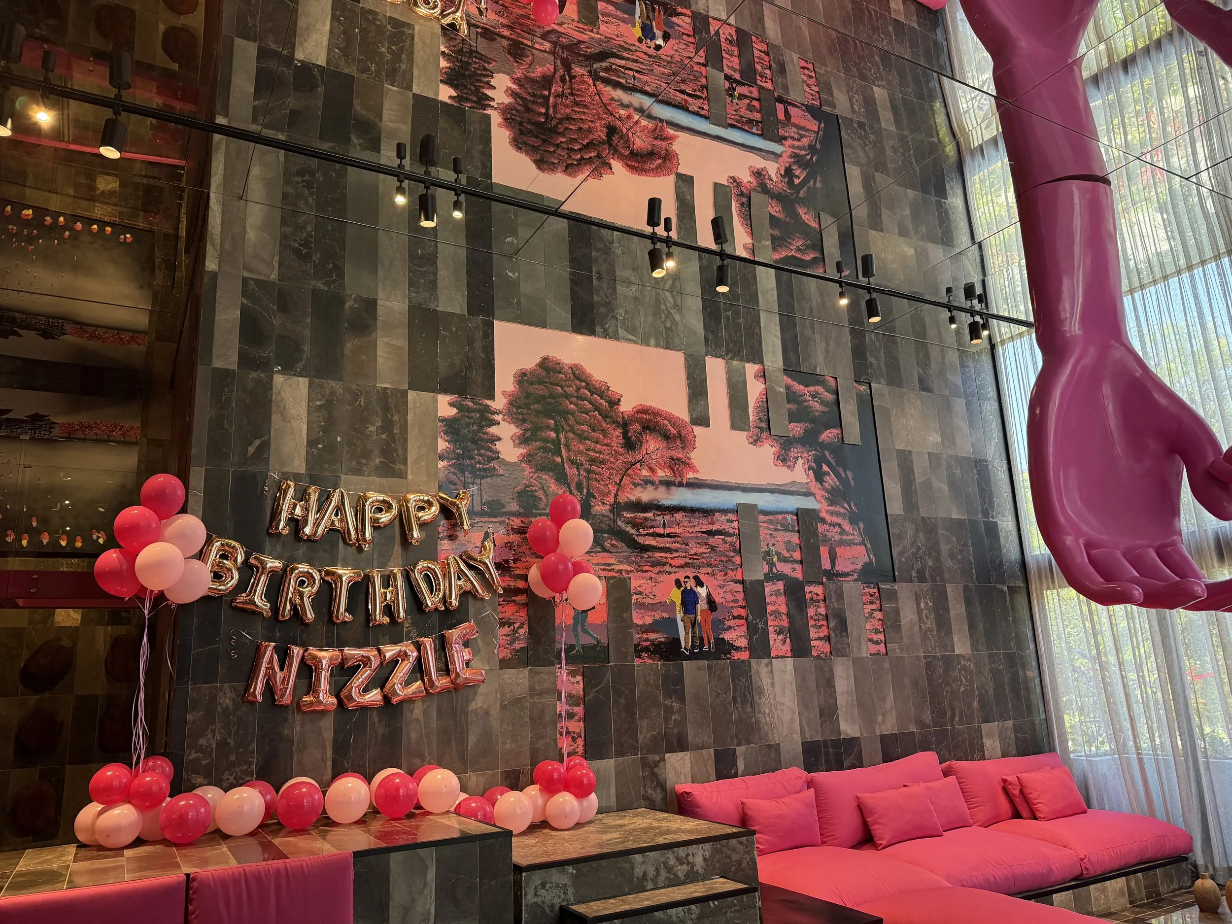 Decorations for a birthday party featuring pink and red balloons, a 'Happy Birthday Nizzle' banner, pink seating, and artwork of trees and a couple on the wall.