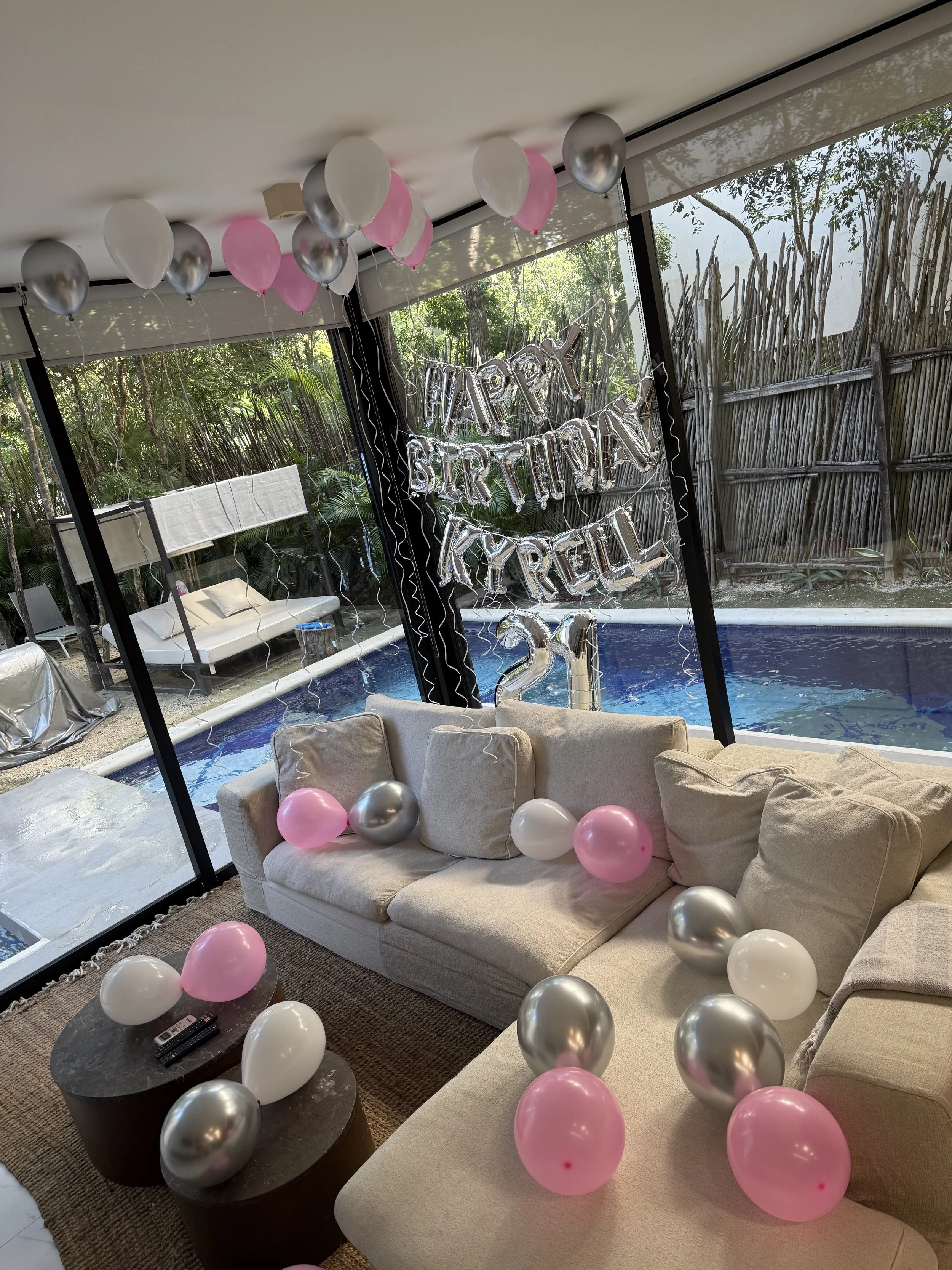 Indoor living room decorated with pink, white, and silver balloons, with a glass wall showing a backyard pool, and a "Happy Birthday" balloon banner.