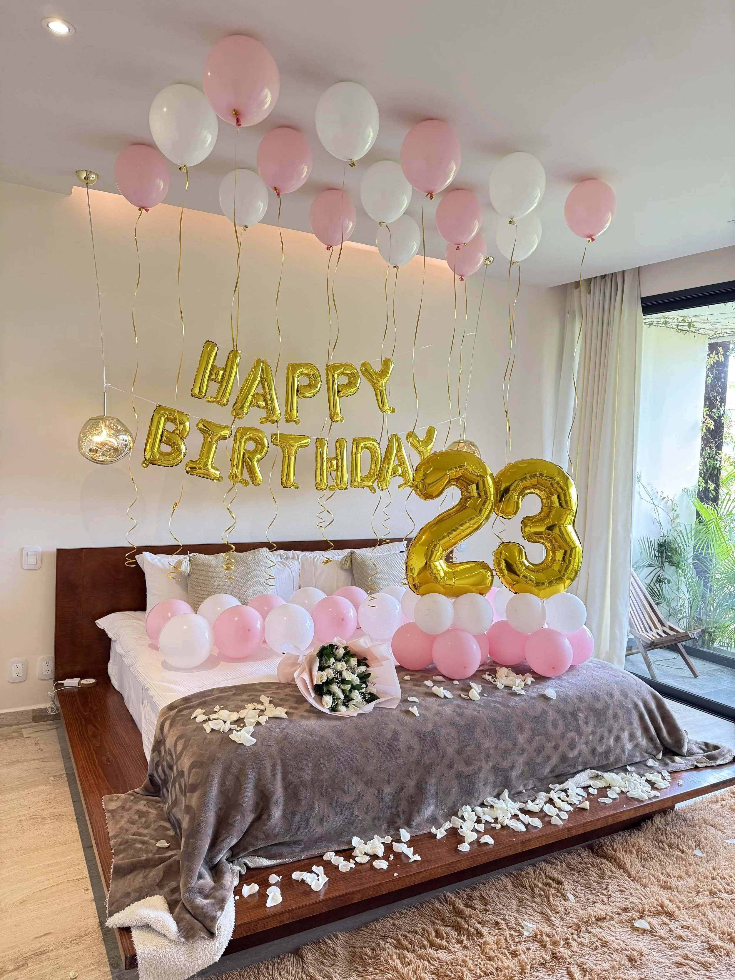 A bedroom decorated for a birthday celebration with pink and white balloons, gold letter balloons spelling 'HAPPY BIRTHDAY', and large gold number balloons forming '23'. A bouquet of white roses on the bed, with flower petals scattered on the bed and