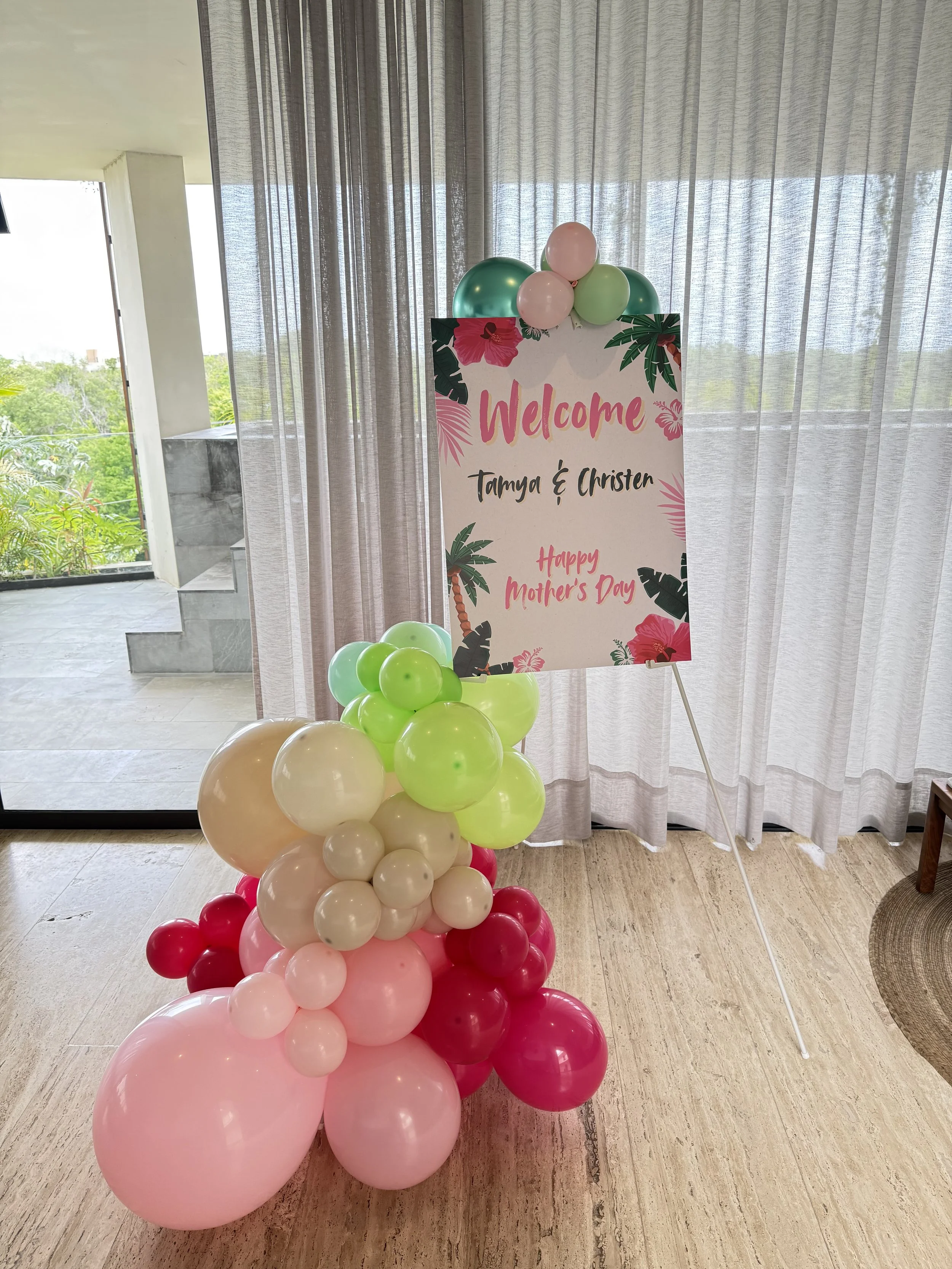 Decorative sign with pink, green, and cream balloons at a tropical-themed Mother's Day celebration. The sign reads: "Welcome Tammy & Christen Happy Mother's Day" and is decorated with large tropical leaves and hibiscus flowers.