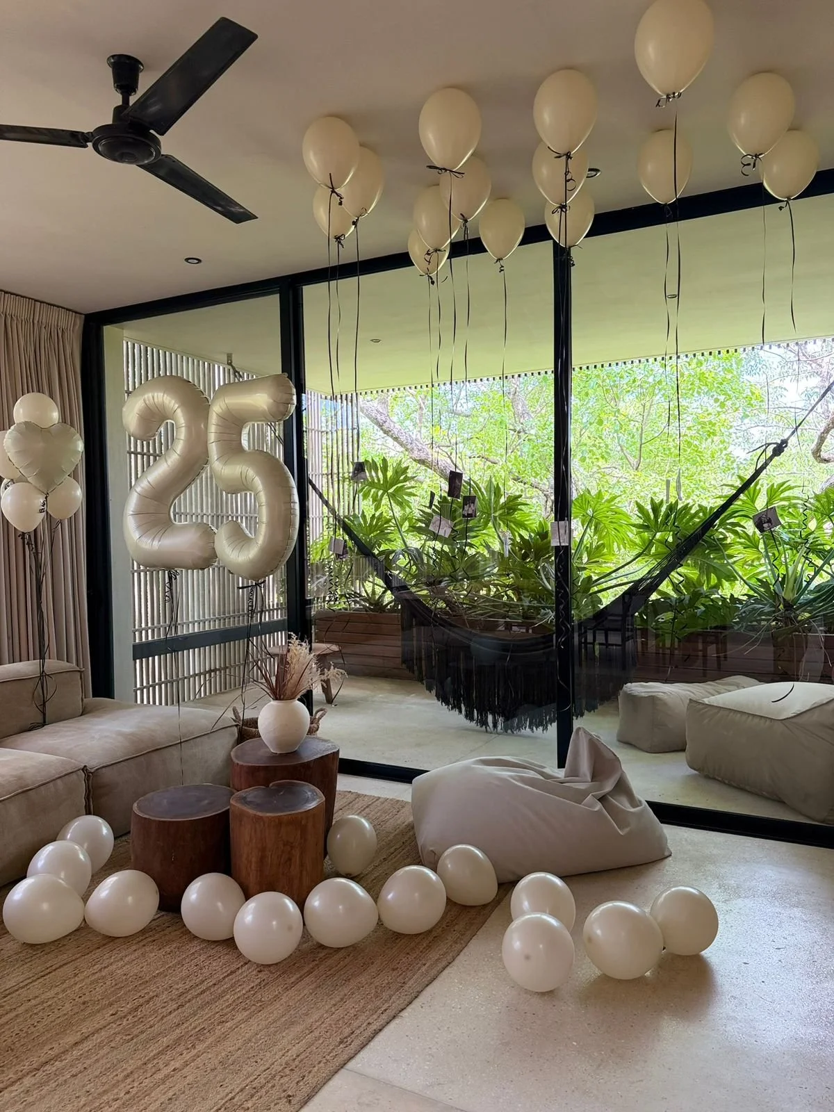 Living room decorated with white balloons, including a large balloon shaped as the number 25, and a black hammock outside near greenery.
