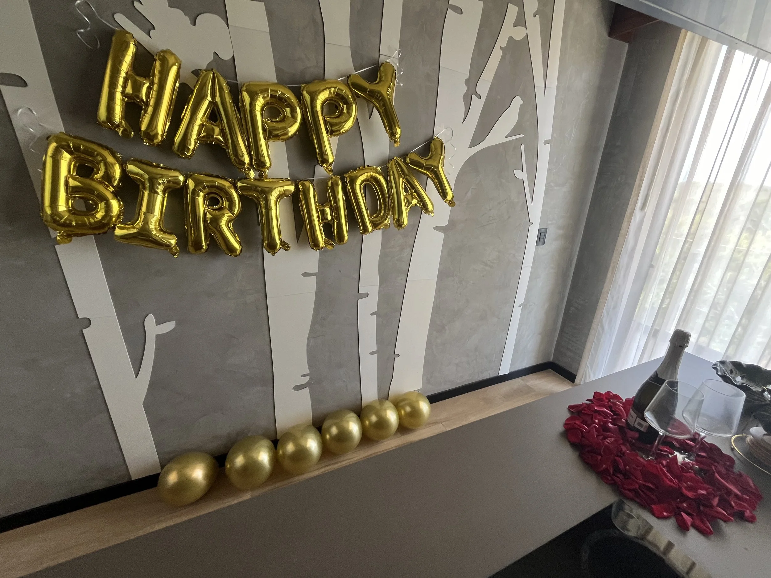 Gold balloons spell out 'Happy Birthday' on a gray wall with a tree silhouette, with gold balloons at the base, and a table with a bottle, glasses, and a red rose petal arrangement.