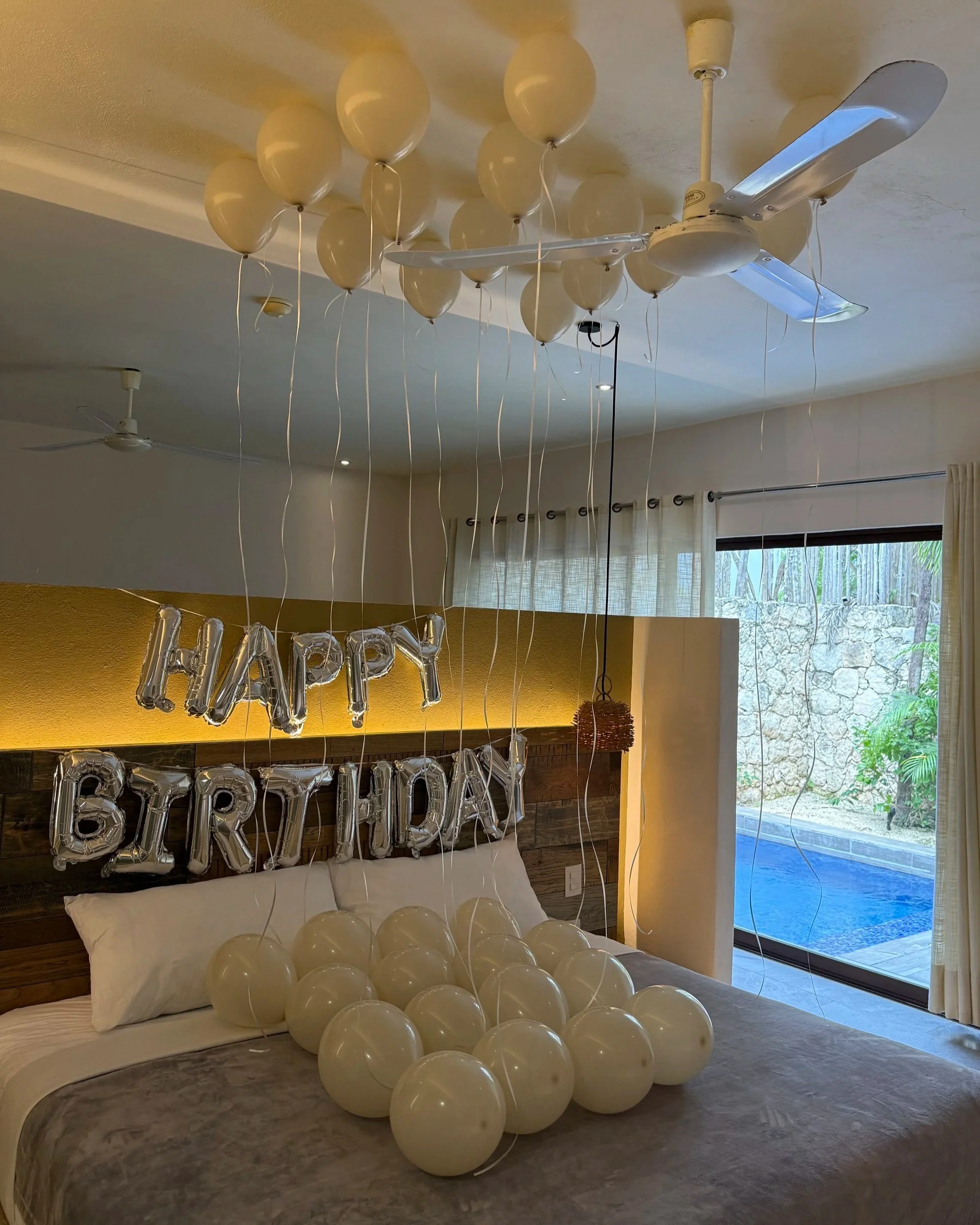 Decorated bedroom with a 'Happy Birthday' silver balloon banner, cream balloons on the bed and attached to the ceiling, and white balloons hanging from the ceiling, with window showing outdoor pool and garden.