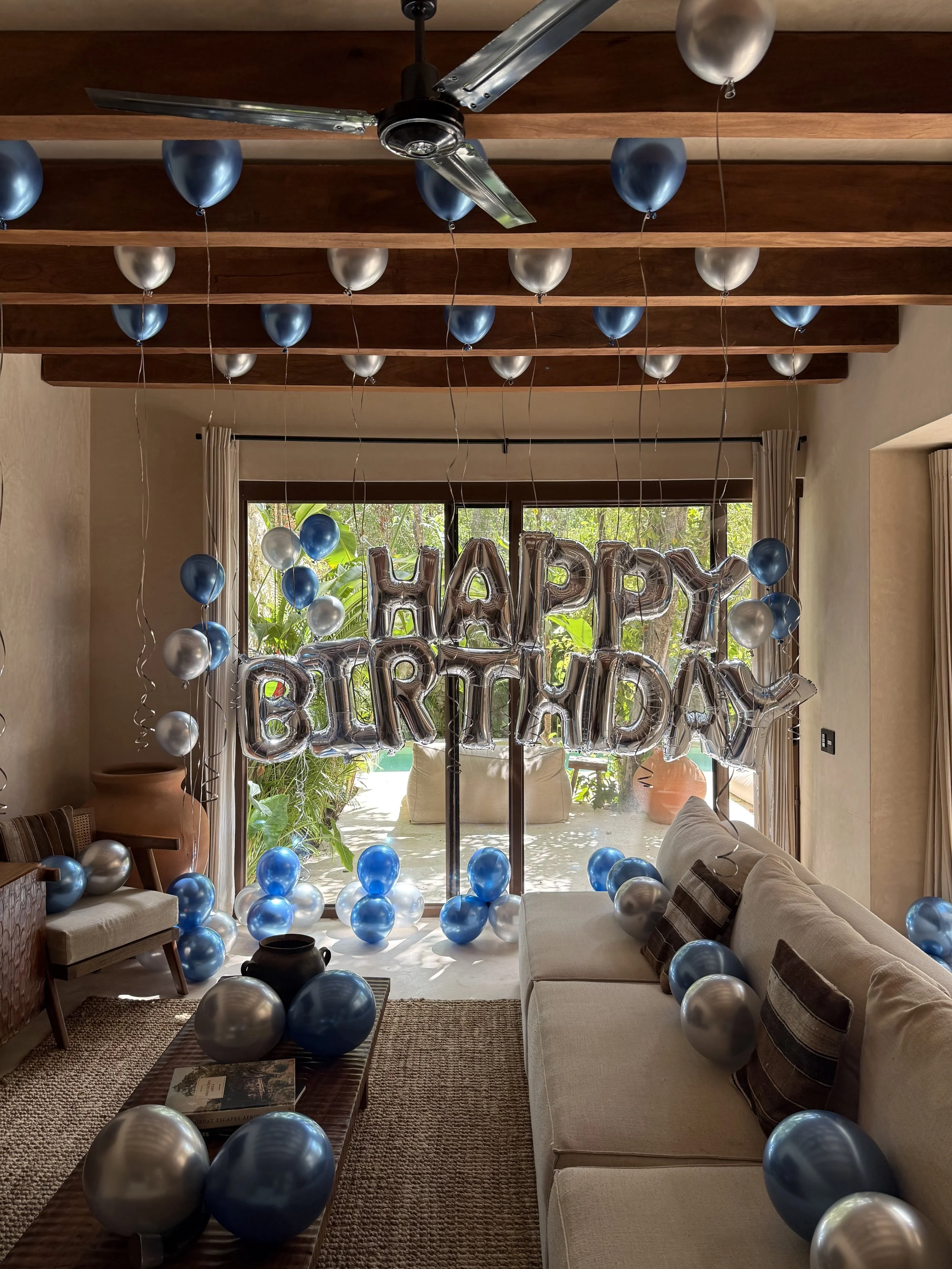Living room decorated with blue and silver balloons, silver foil "Happy Birthday" balloons, a window with greenery outside, a ceiling fan, and a coffee table with books.