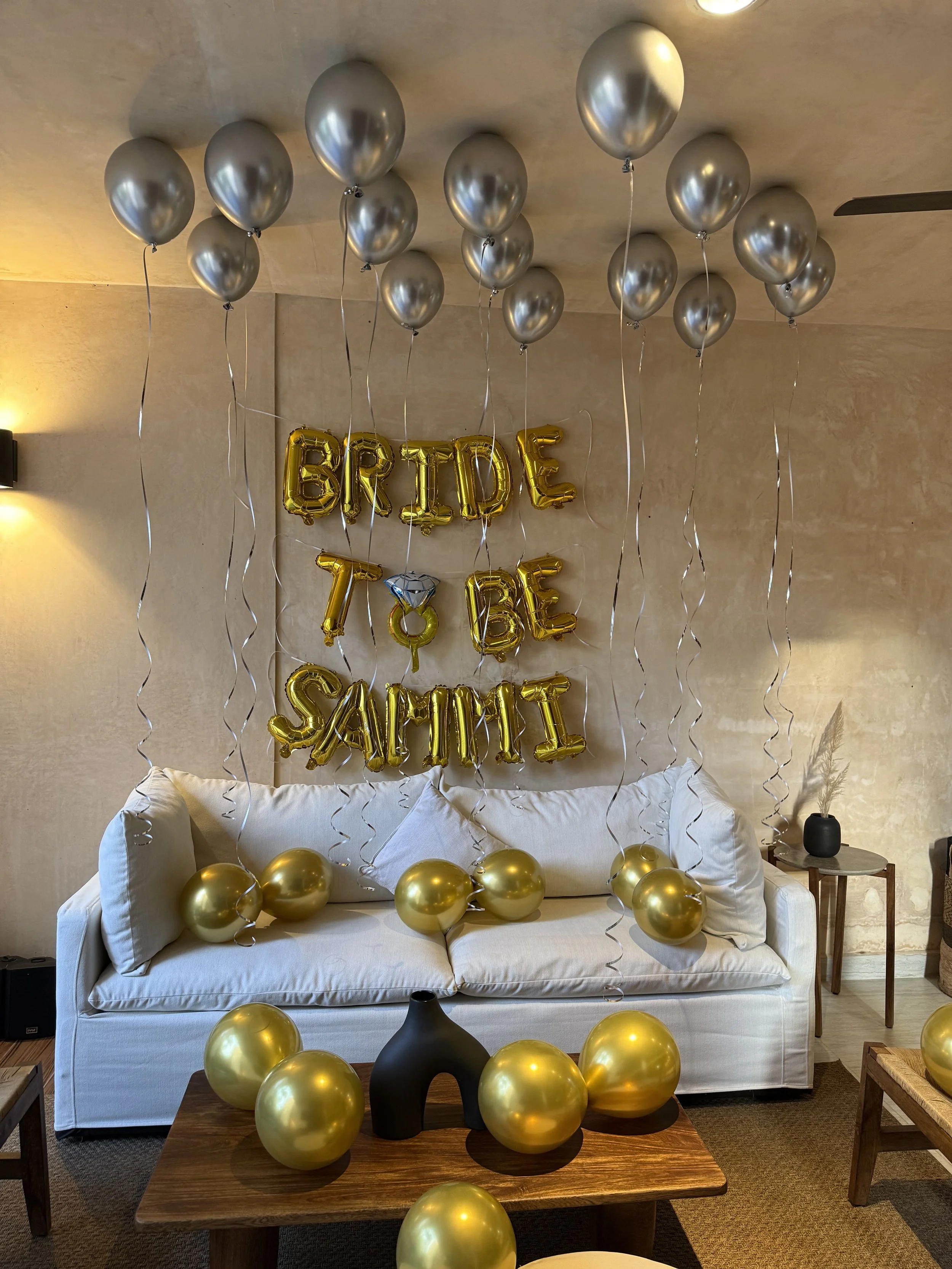 Decorations for a bridal shower with gold and silver balloons, including gold balloon letters spelling 'BRIDE TO BE SAMMI', hanging from the ceiling, with some balloons on the couch and table.