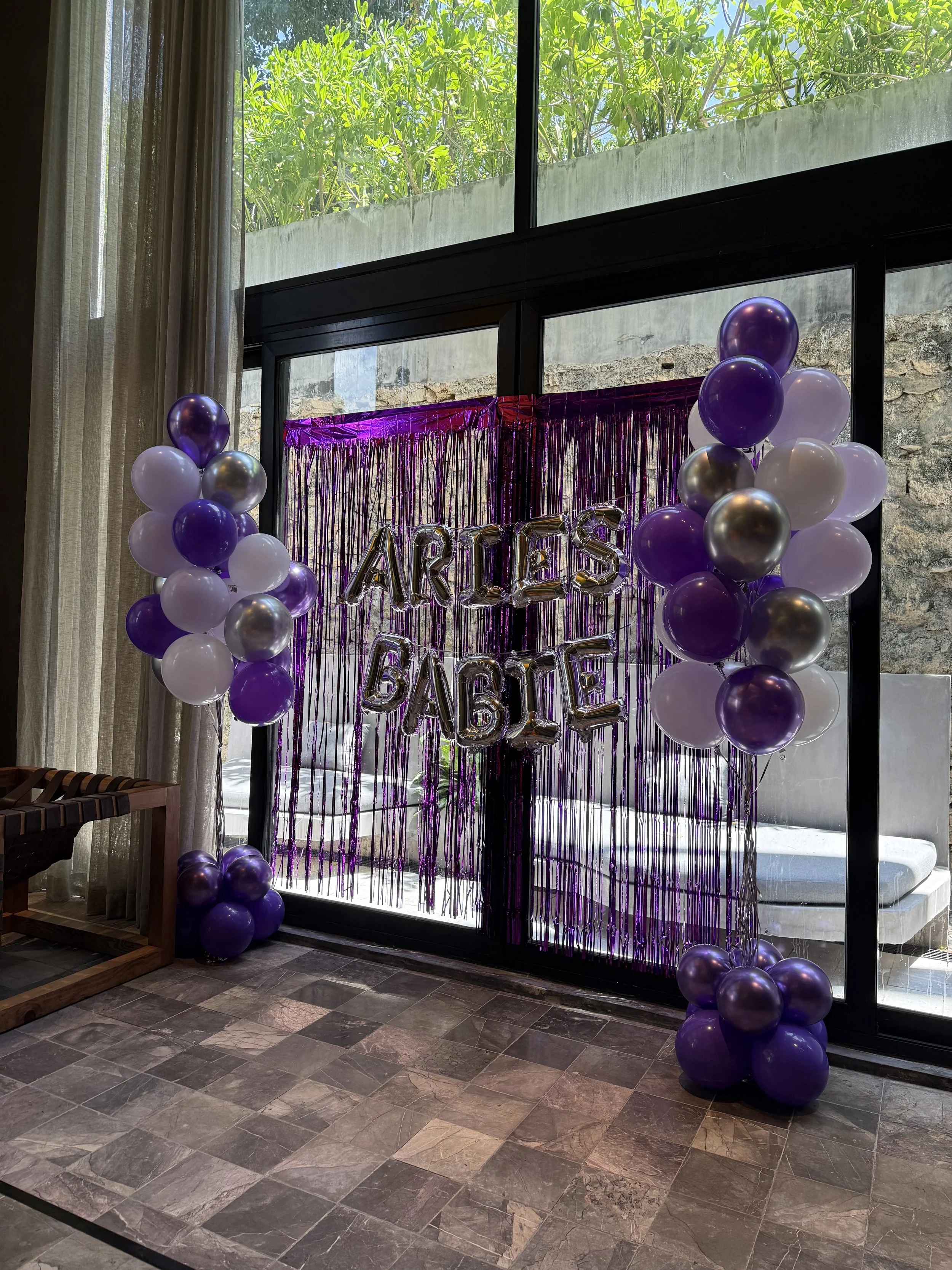 Indoor decoration with purple and white balloons, purple tinsel backdrop, and metallic balloons spelling out 'ARTES' and 'BAILE' in front of a glass door with a tree outside.