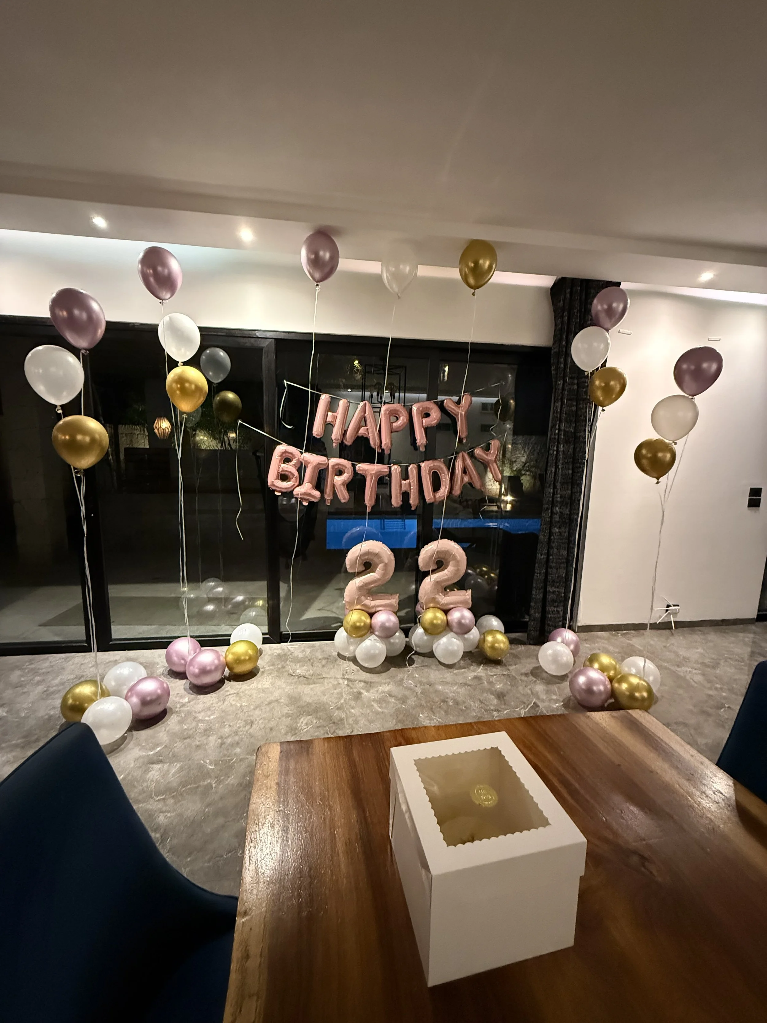Decorated room with pink, white, gold, and silver balloons, including a large 'Happy Birthday' balloon banner and a birthday number '22' balloon, set against a sliding glass door and dark curtains.