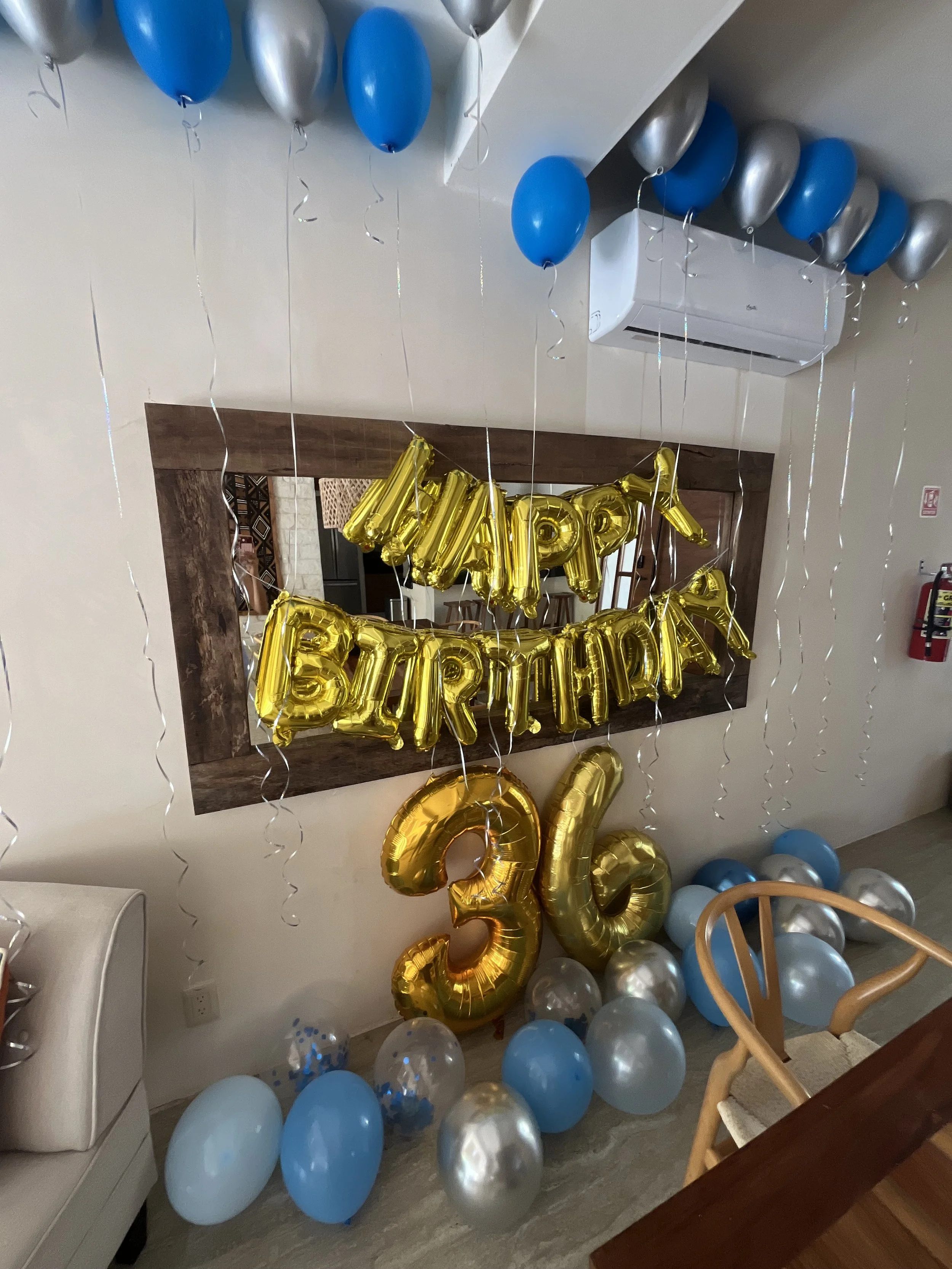 Decorated party area with blue, silver, and gold balloons, a large gold '36' balloon, and a gold 'Happy Birthday' balloon banner reflected in a mirror.