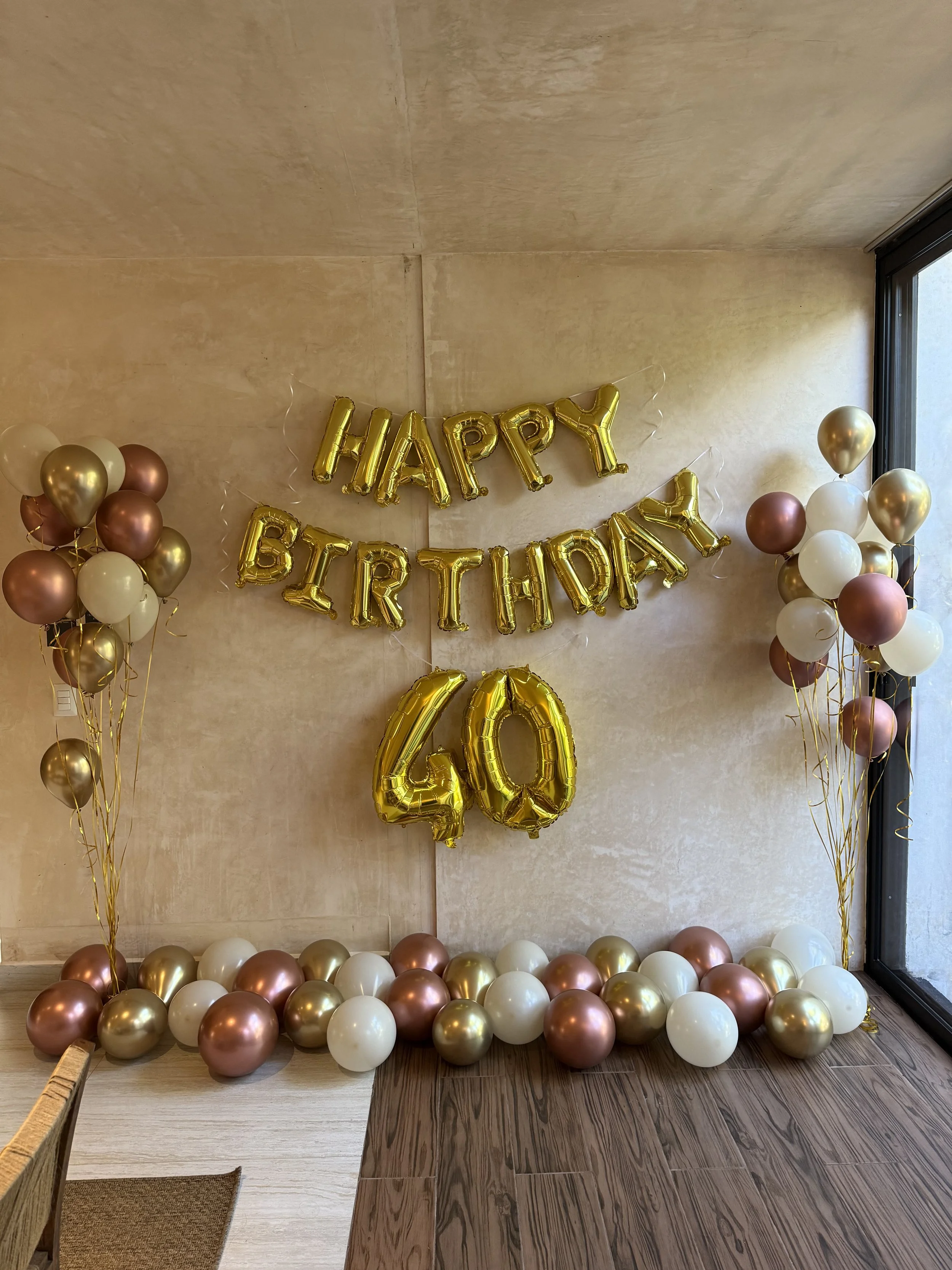 Decorations for a 40th birthday celebration including gold, white, and rose gold balloons, with a banner that reads 'HAPPY BIRTHDAY 40' in gold foil balloons, against a beige wall near a window.