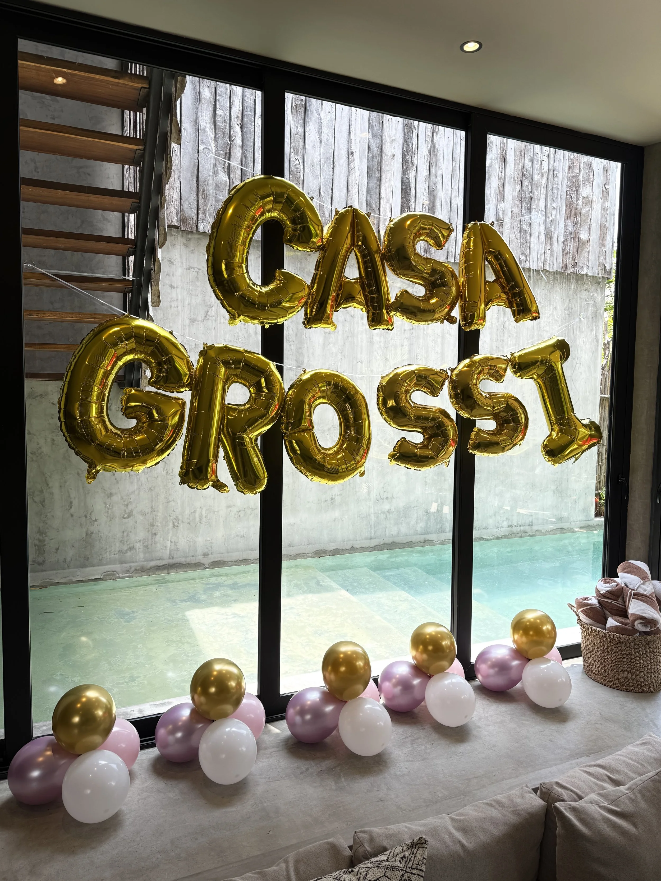 Golden balloon letters spelling 'CASS' hanging in front of large glass doors, with pink, white, and gold balloons lining the floor below.