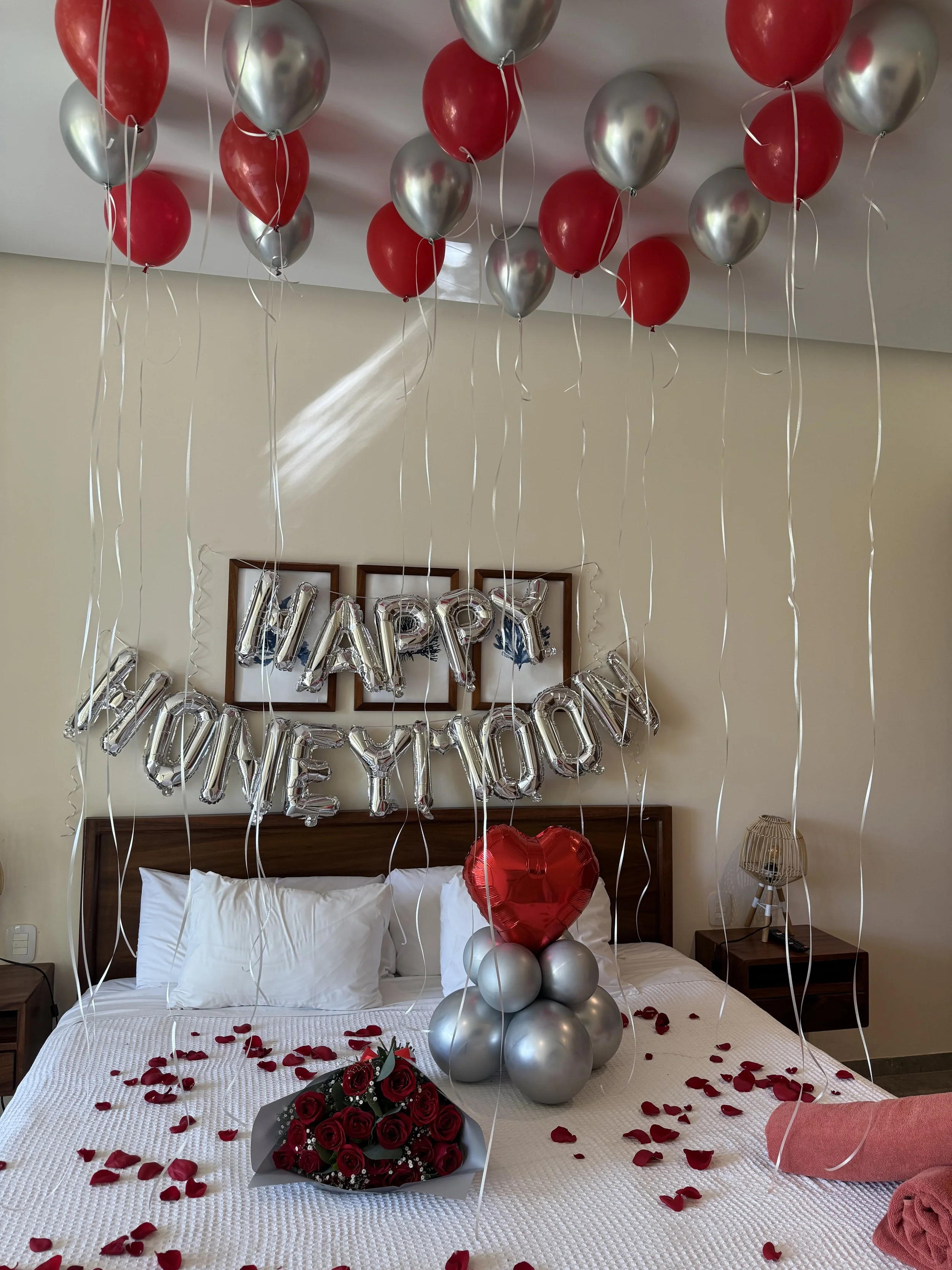 A decorated hotel room bed with red and silver balloons, a bouquet of red roses, scattered rose petals, and silver foil balloons spelling 'HAPPY HONEY MOON'.