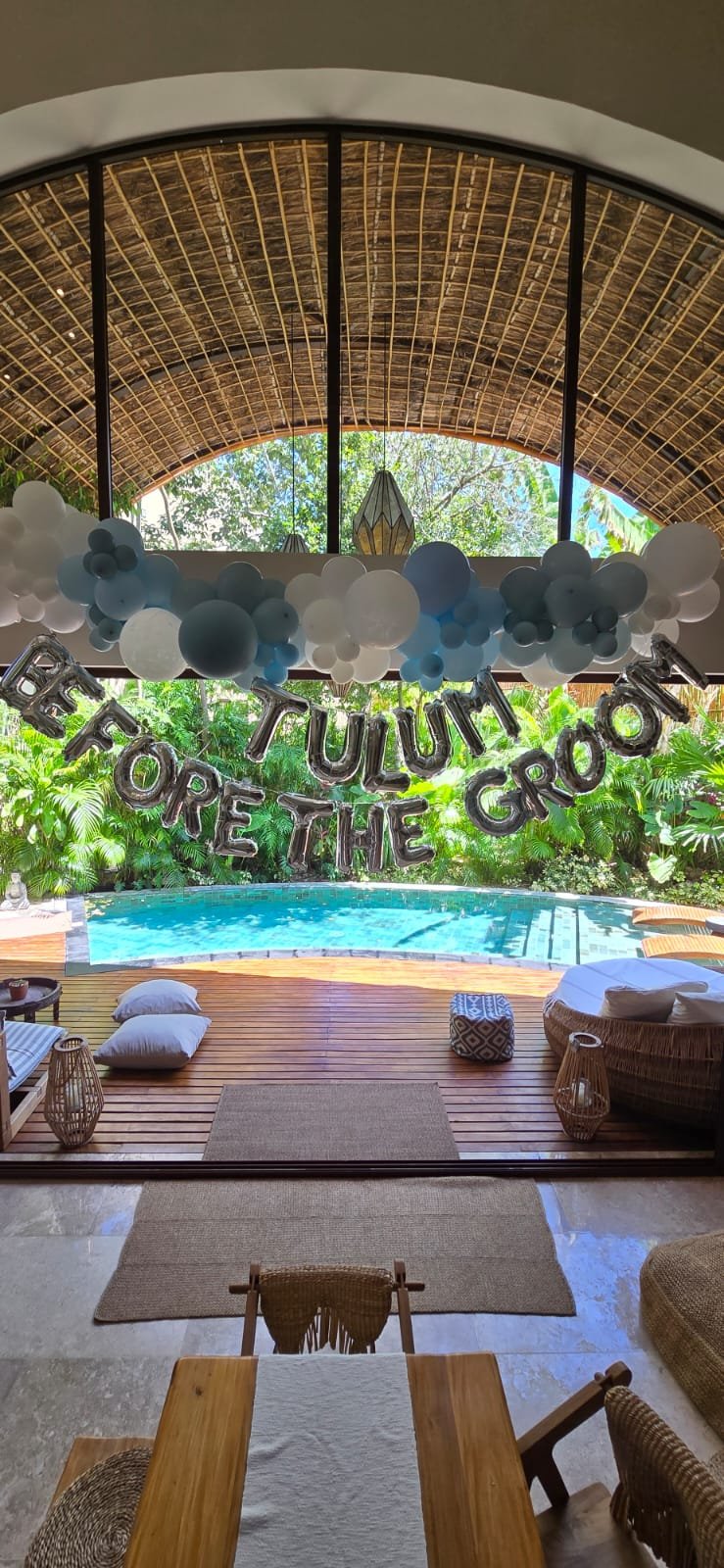 Indoor space decorated for a celebration with a banner that says TULUM BEFORE THE GROOM, surrounded by balloons and a view of a pool and lush greenery outside.