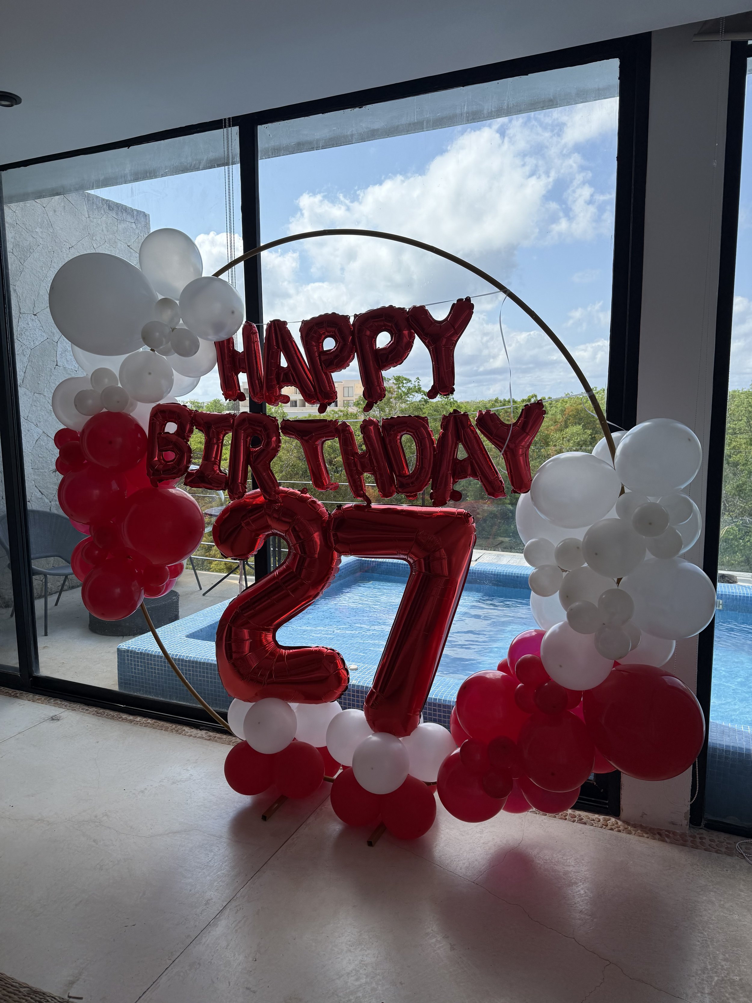 Colorful balloon arrangement for a birthday celebration, featuring a large red number '27' balloon in the center, with white and red balloons surrounding it, set indoors near a window with a view of the sky and trees outside.