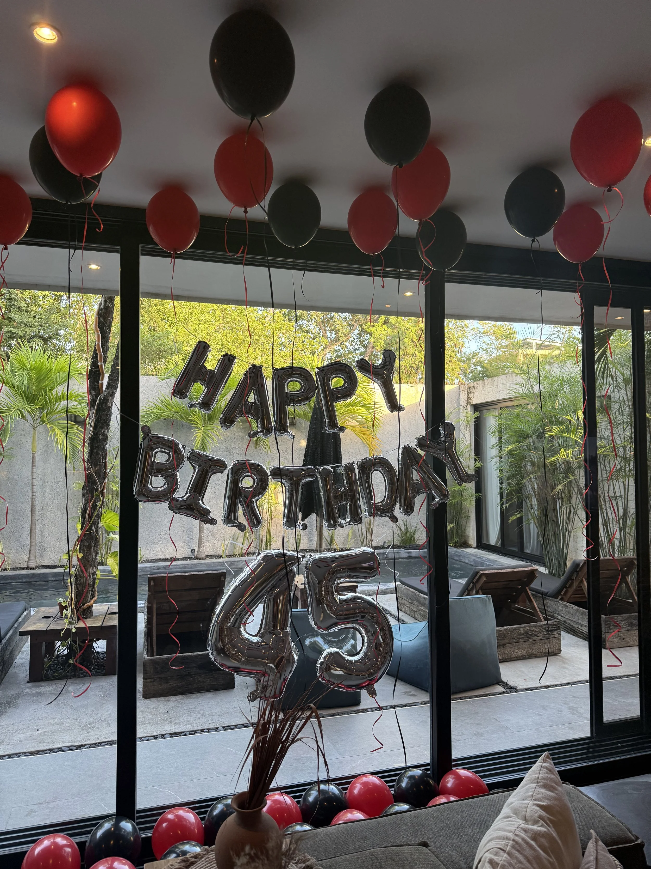 Decorations for a 45th birthday celebration inside a house or apartment, including red and black balloons, a "Happy Birthday" banner in black balloons, and a large silver number "45" balloon. The decorations are set against a glass window or sliding 