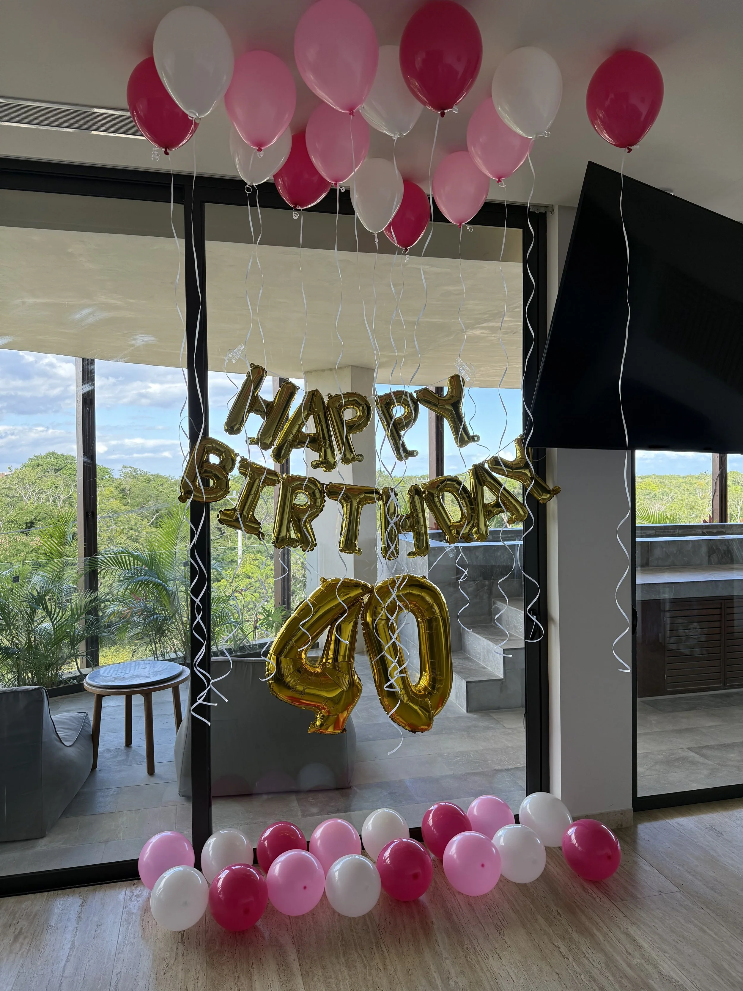 Decorations for a 40th birthday celebration with pink and white balloons, gold number balloons '40', and a 'Happy Birthday' foil banner, set in a room with glass doors and a view of greenery outside.