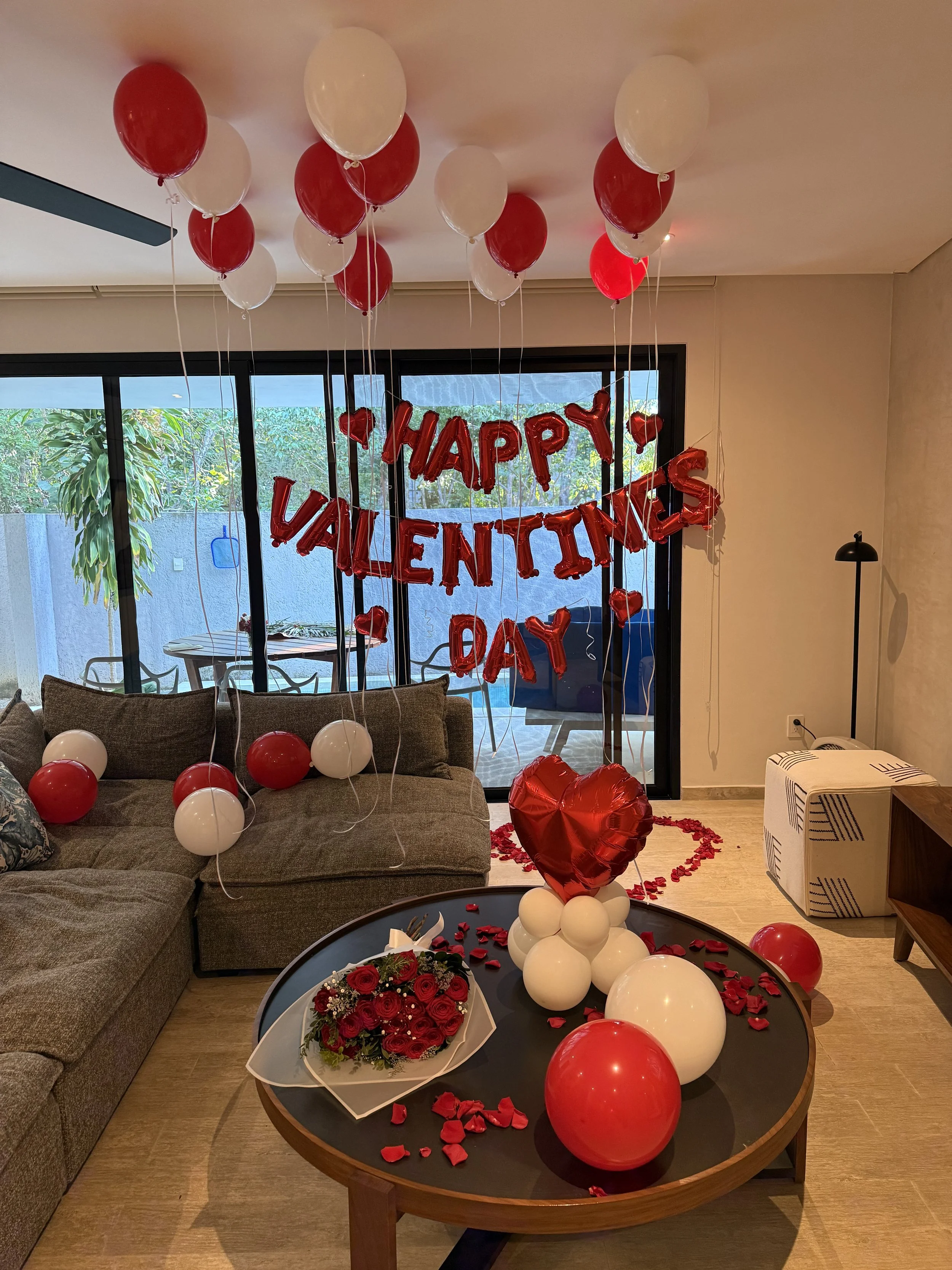 Valentine's Day decorations in a living room with red and white balloons, a heart-shaped balloon arrangement, a floral bouquet, and a 'Happy Valentine's Day' banner.
