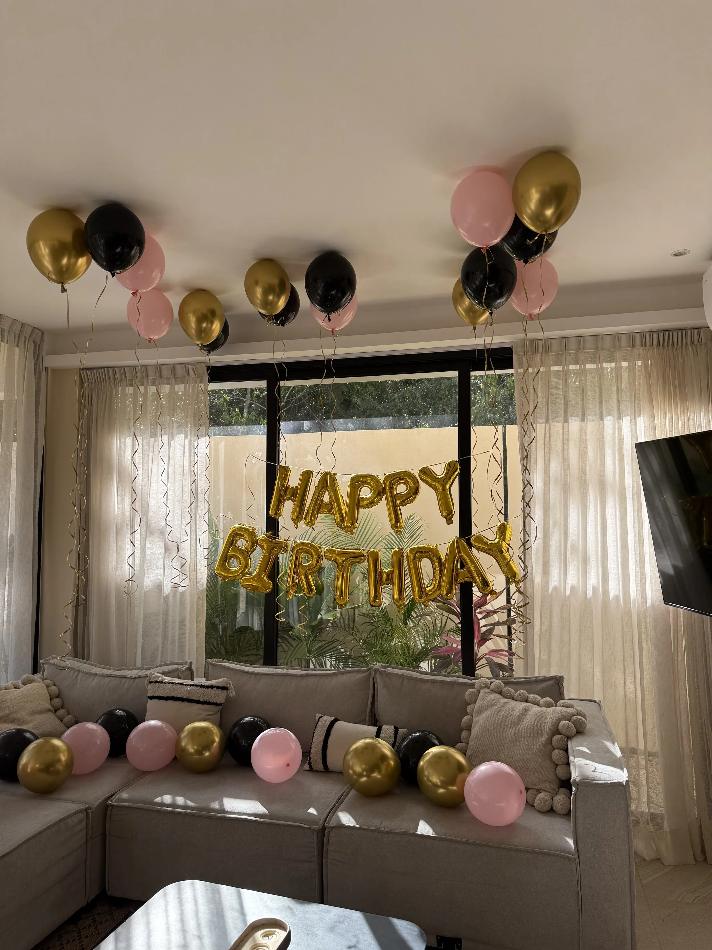 A living room decorated for a birthday celebration with pink, black, and gold balloons, a gold 'HAPPY BIRTHDAY' banner, and a large window with sheer curtains.