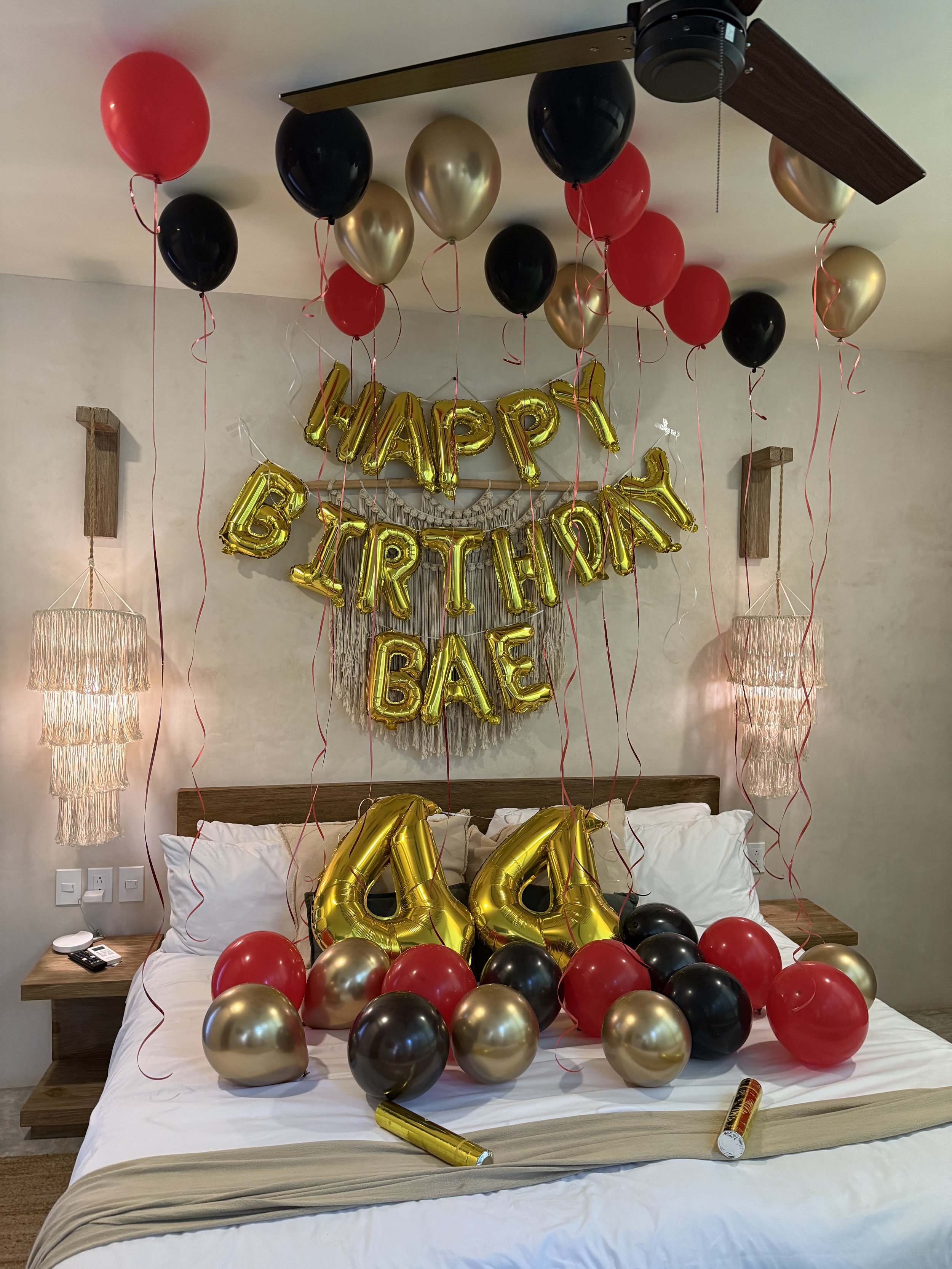 Decorated celebration birthday on a bed with balloons, including black, red, and gold helium balloons, gold foil 'happy birthday' balloons, and large gold number balloons '44'. A background banner spells out the name 'Bae' in gold letters. The room h
