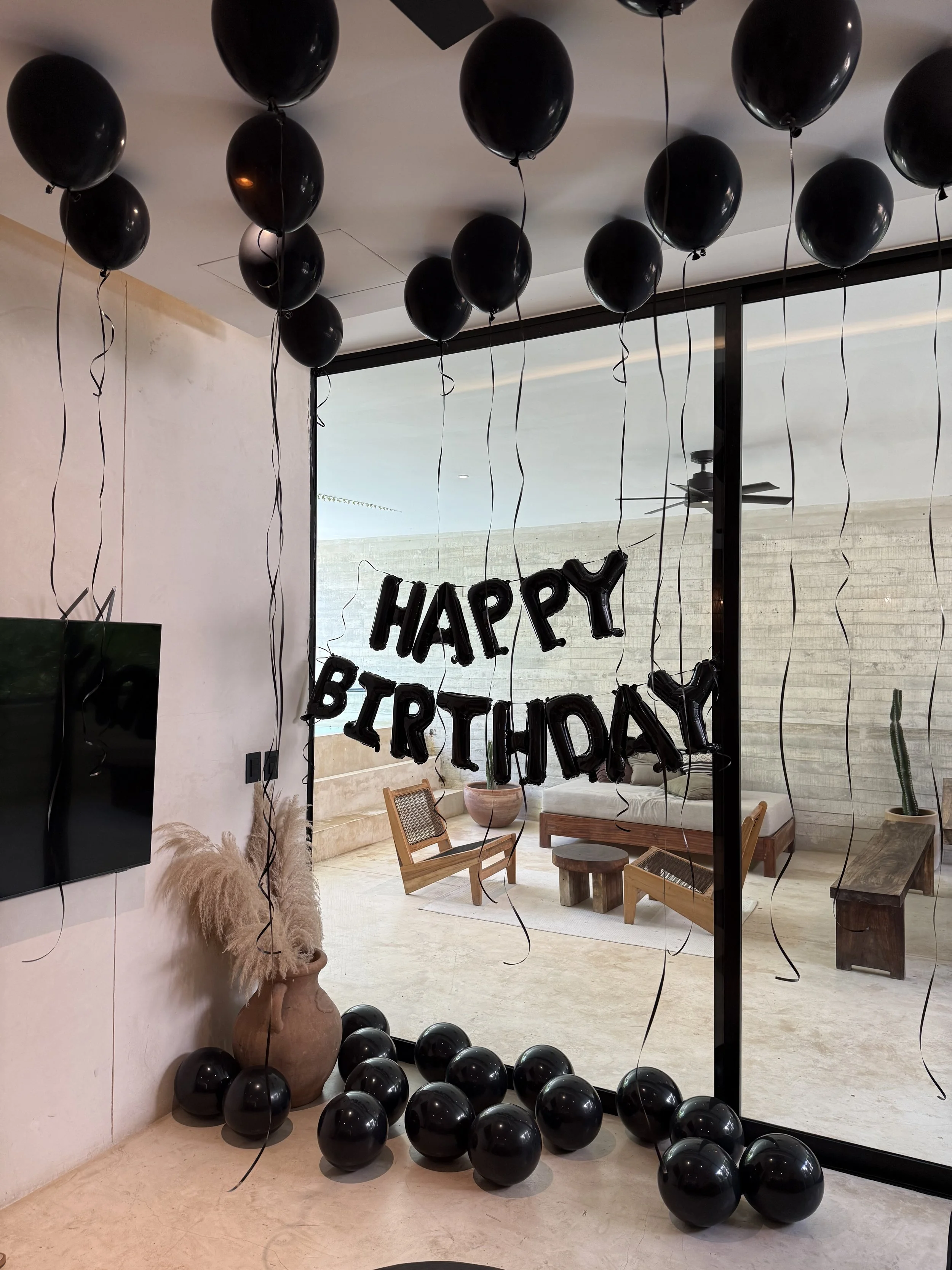 Decorative black balloons hanging from ceiling, with some on the floor, in front of a large window. A "Happy Birthday" banner is visible on the window.