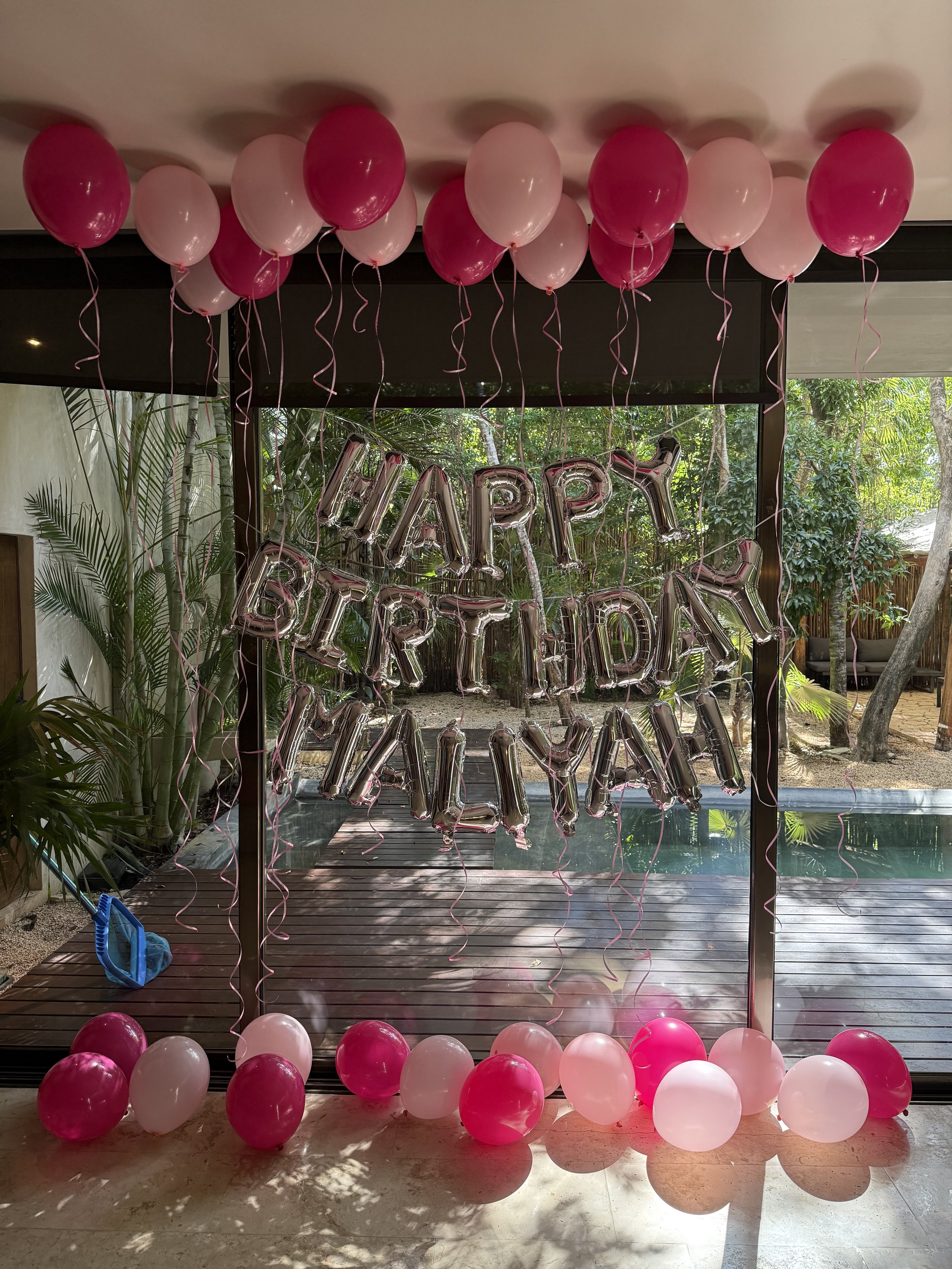 Decorative balloons in pink and white with a 'Happy Birthday Melylah' metallic balloon banner in the background, set indoors with large glass windows and greenery outside.