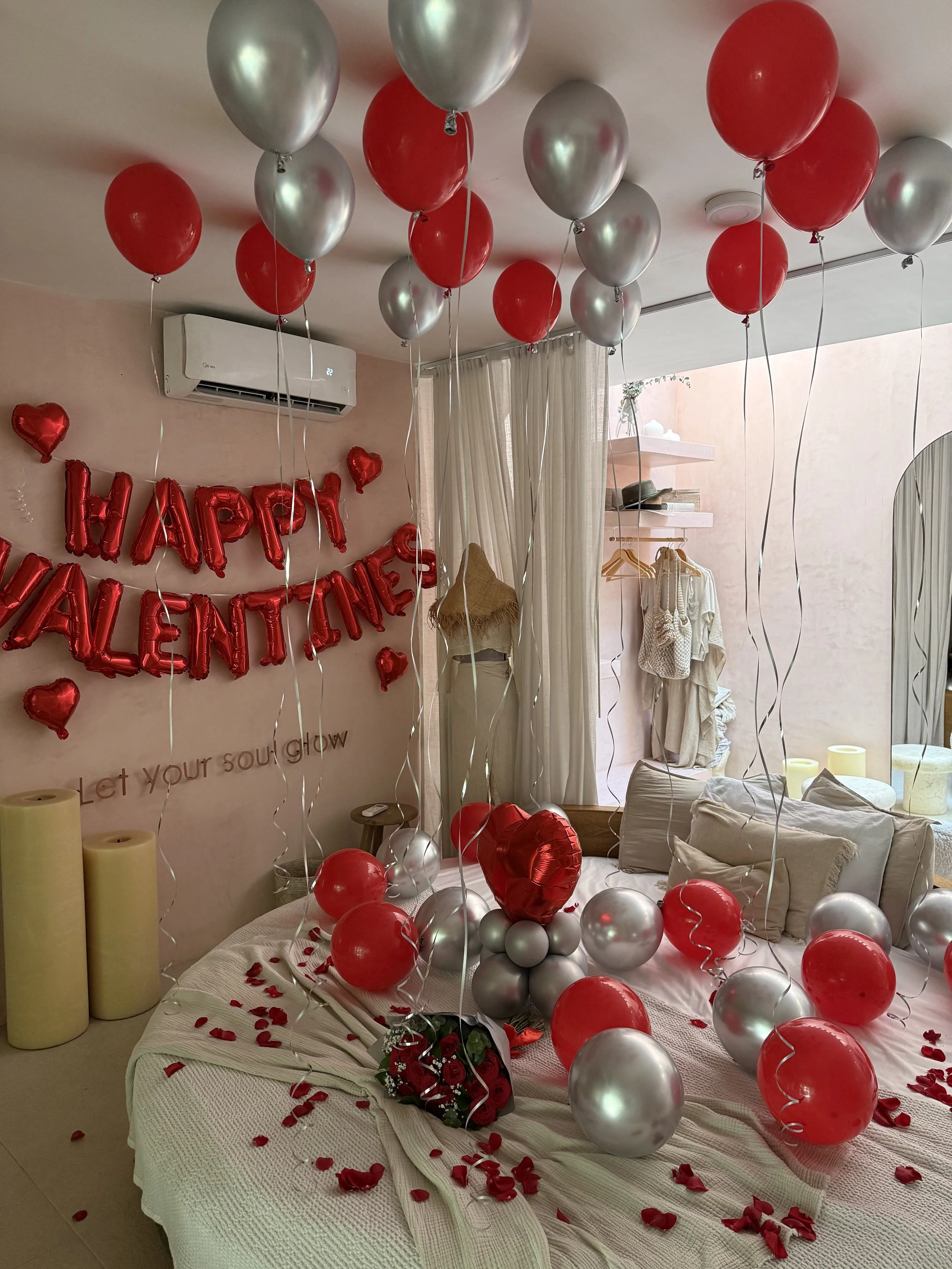 Room decorated for Valentine's Day with red and silver balloons, red heart-shaped balloons, rose bouquet, and a wall display that says 'Happy Valentine's' with hearts and the phrase 'Let your soul glow'.