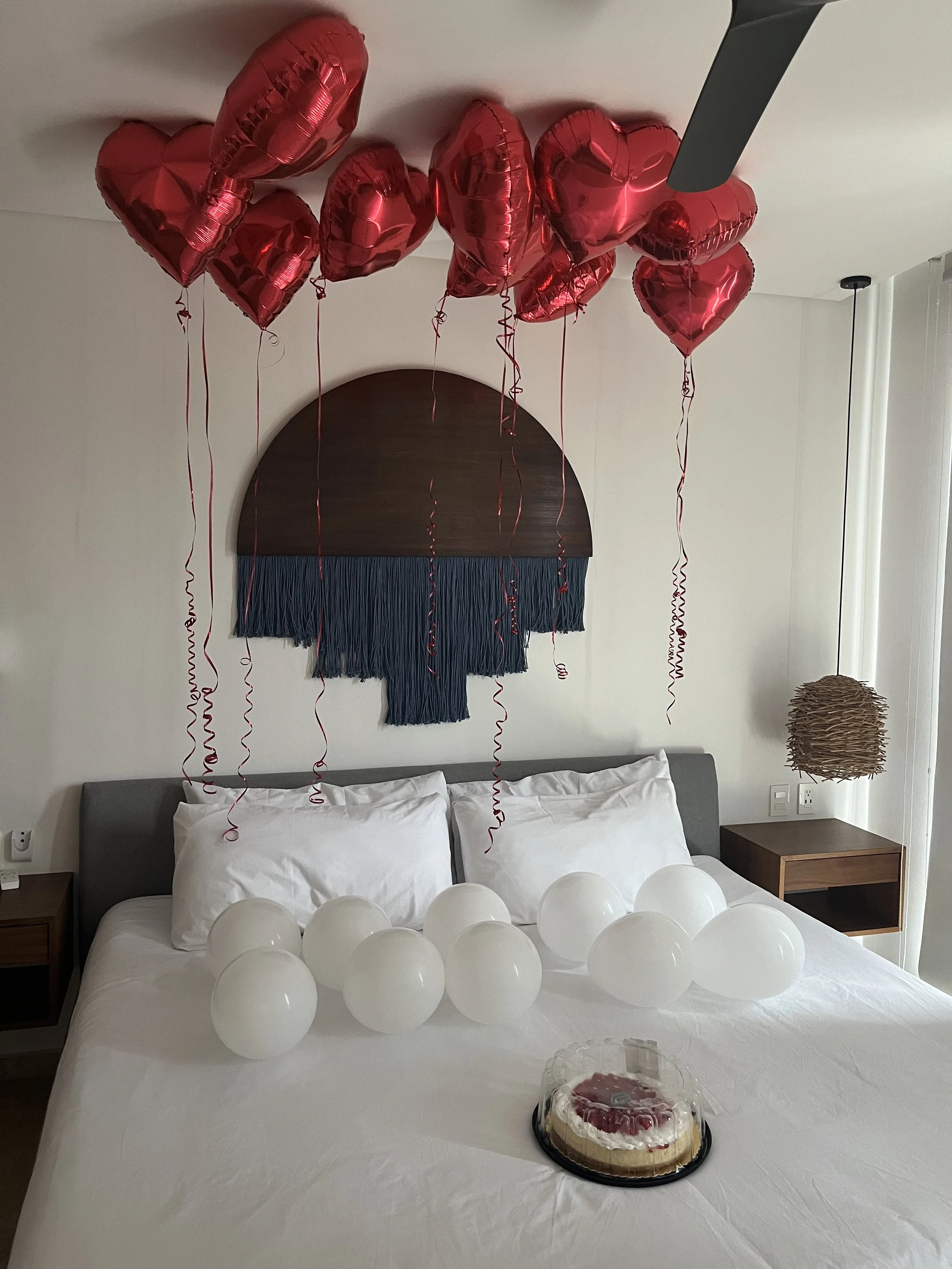 Decorated hotel room with white balloons on bed, a cake, and red heart-shaped balloons attached to the ceiling.