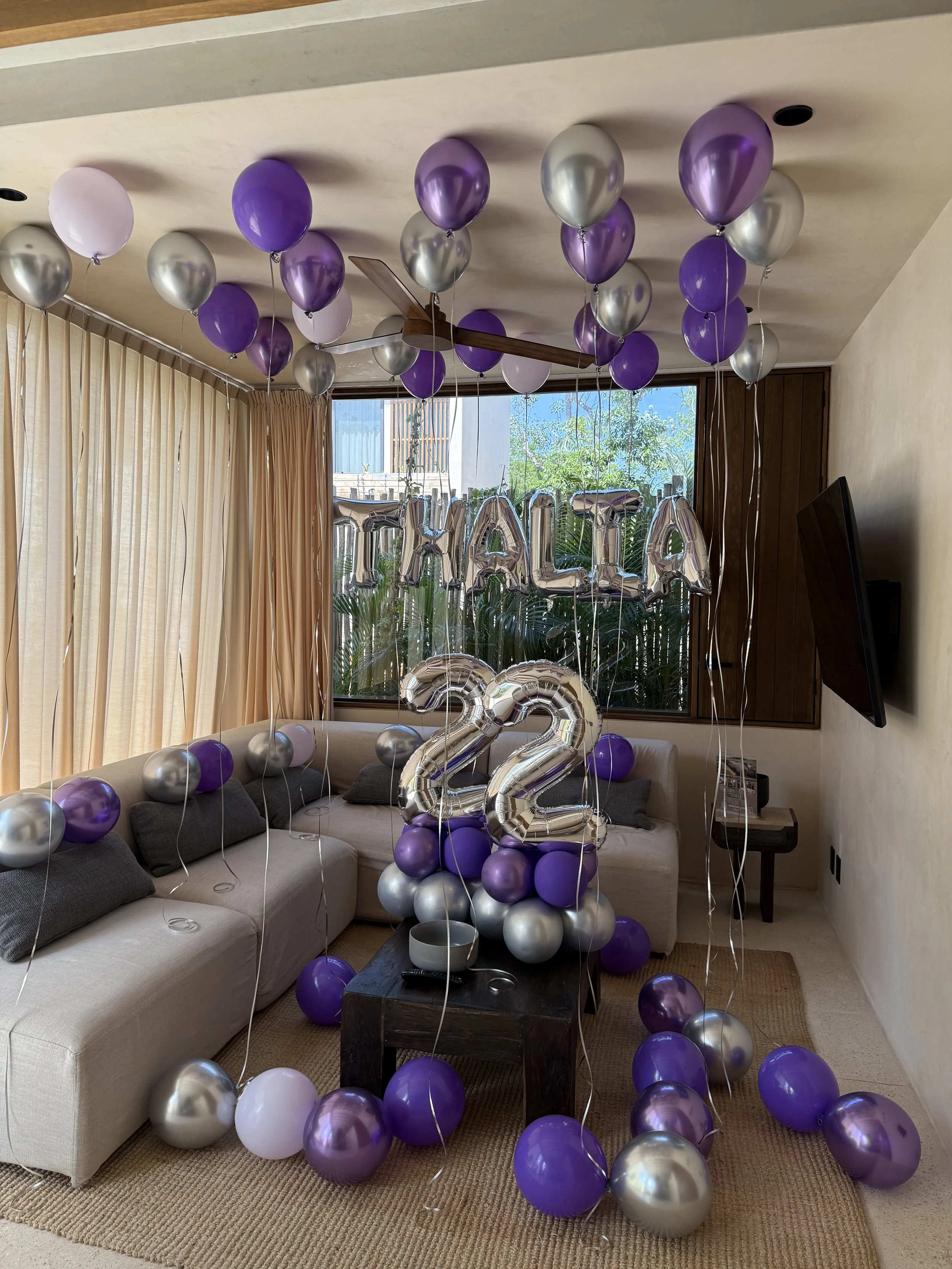 Living room with purple and silver balloons, including a large number 22 balloon, and silver balloon letters spelling 'MALIA' hanging behind the sofa. Balloons are scattered on the floor and tied to furniture.
