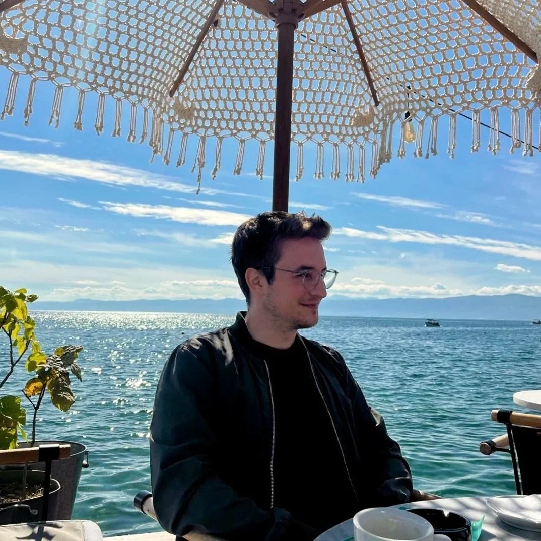 Ein Mann sitzt an einem Tisch im Freien unter einem großen, weißen, gehäkelten Sonnenschirm an einem Wassersee, der wahrscheinlich ein See ist, bei strahlendem Sonnenschein, mit türkisem Wasser, einem blauen Himmel und einigen Wolken.