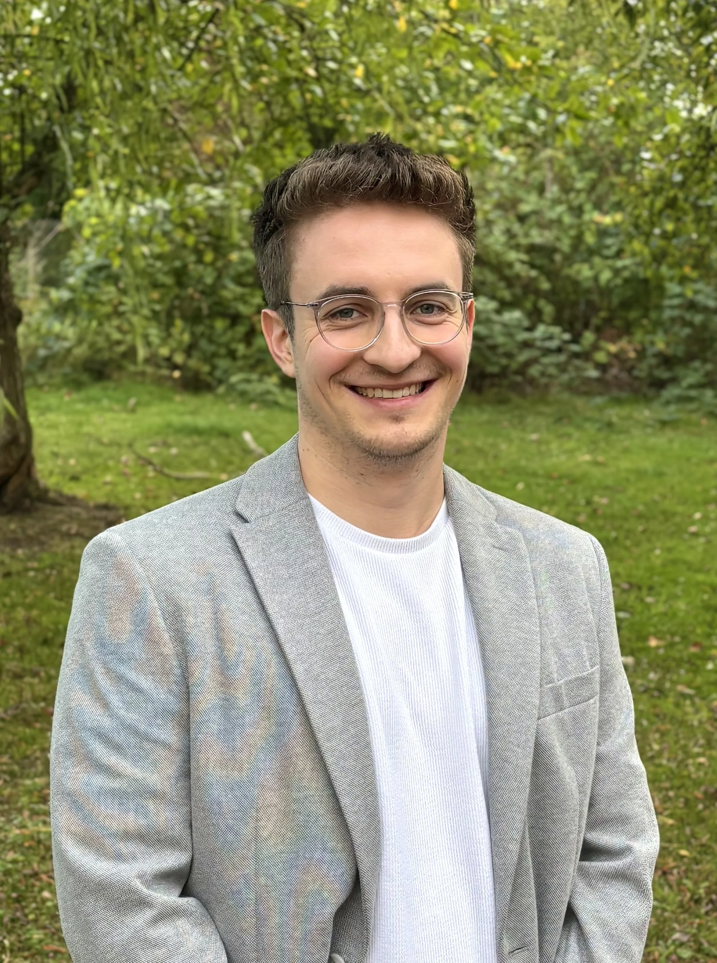 Ein junger Mann mit dunklen Haaren, Brille und einem grauen Anzug steht im Grünen, lächelt und blickt in die Kamera.