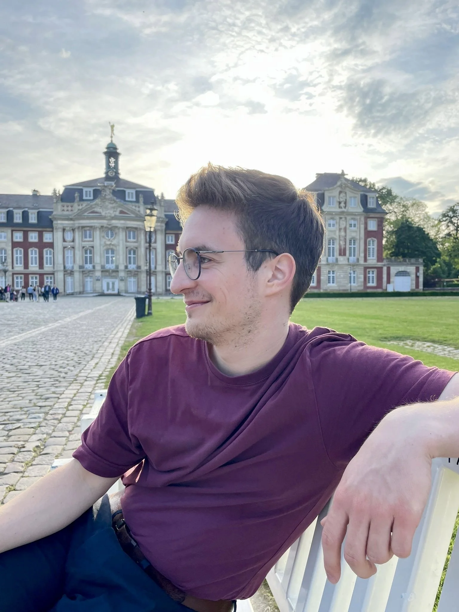 Junge Person mit Brille, sitzt auf einer Bank vor einem Schloss, bei Sonnenuntergang, im Freien.