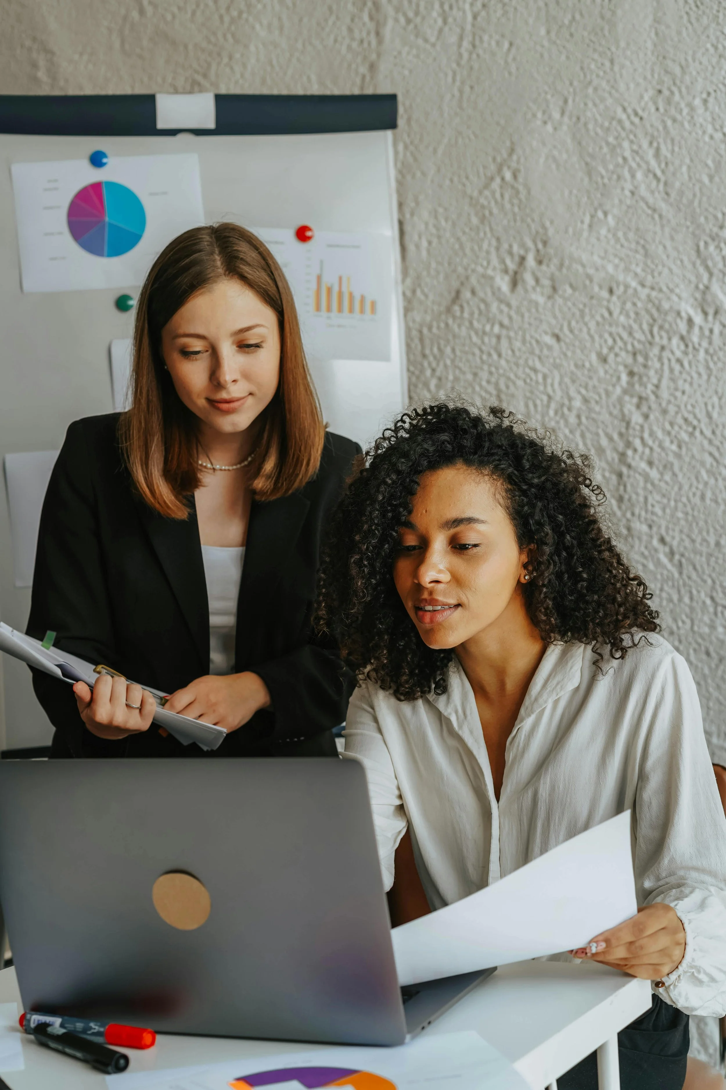 Two women working together in an office, looking at documents and a laptop, with charts on a whiteboard in the background.