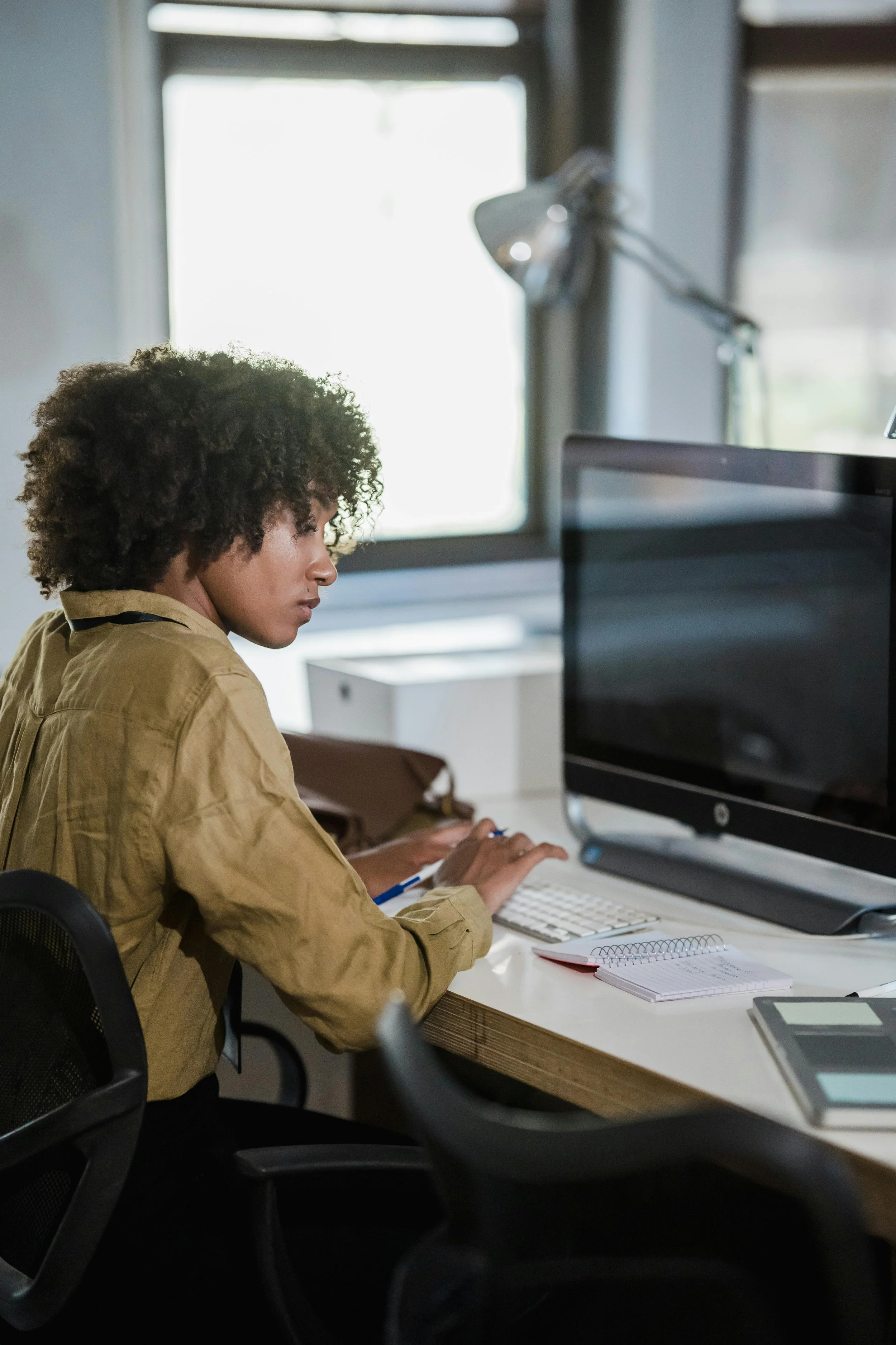 A woman with curly hair sitting at a desk in front of a computer monitor, taking notes with a pen in a bright, modern office space.