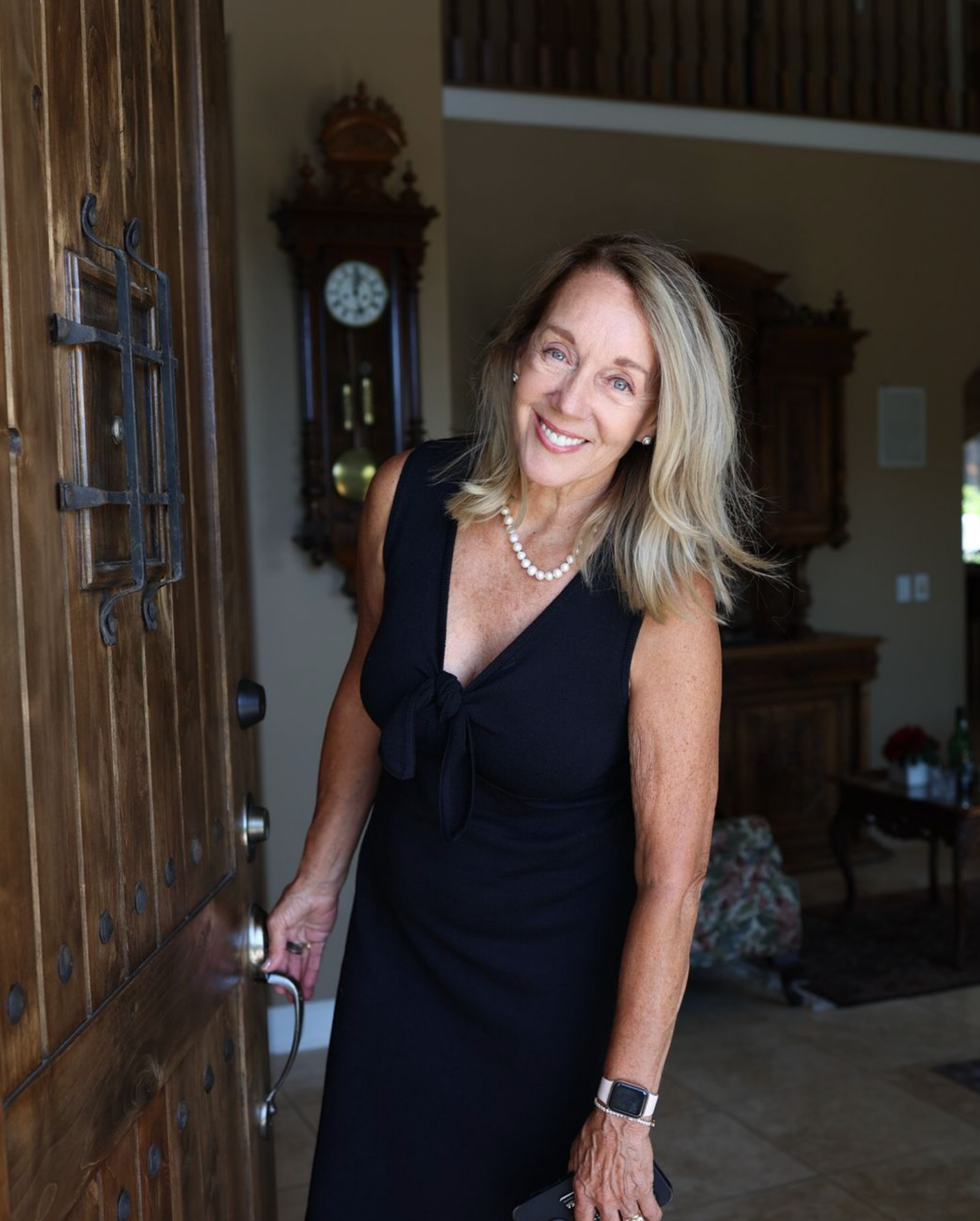 A woman in a black dress standing at a doorway, smiling, with a pearl necklace and a smartwatch.