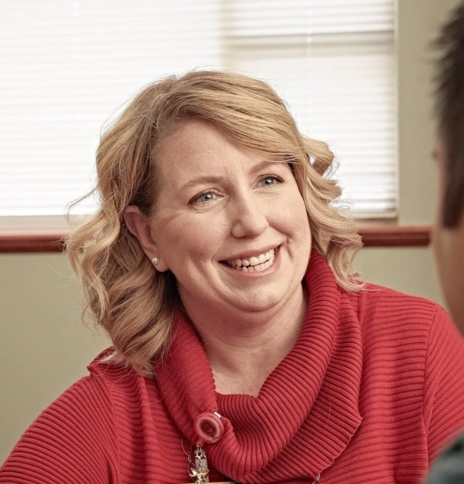 A woman with shoulder-length blonde hair smiling, wearing a red ribbed turtleneck sweater, sitting indoors near a window with blinds.