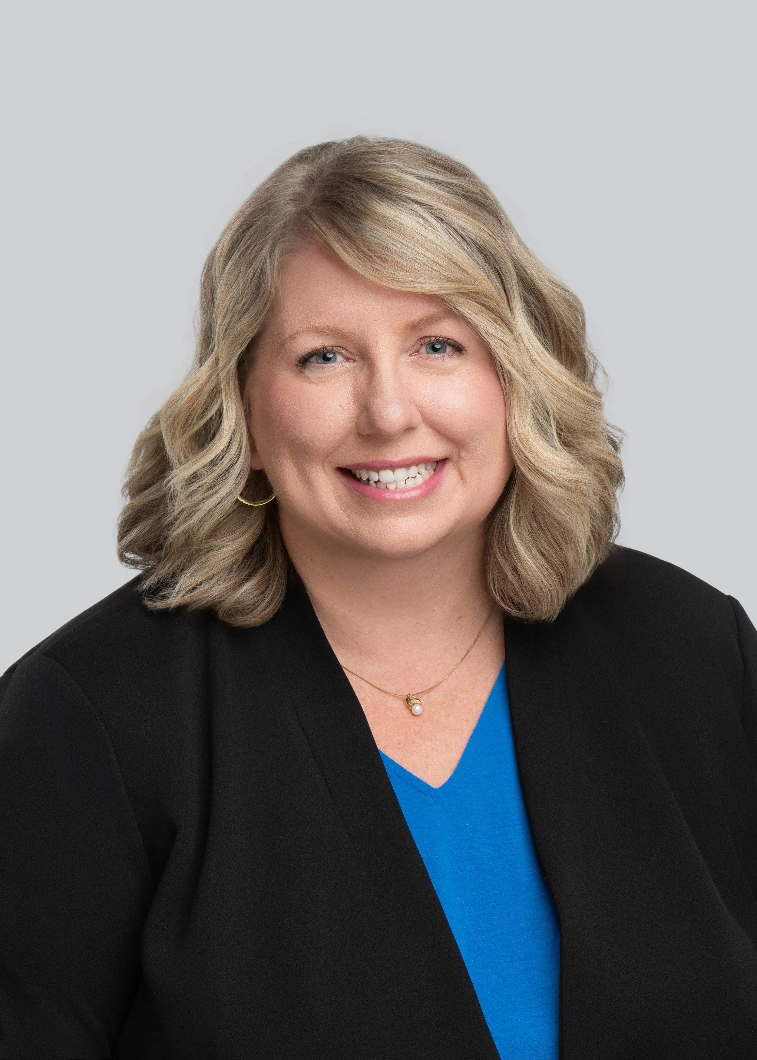 Headshot of a smiling woman with blonde curly hair, wearing a black blazer, a blue top, and a pearl pendant necklace, against a plain light gray background.
