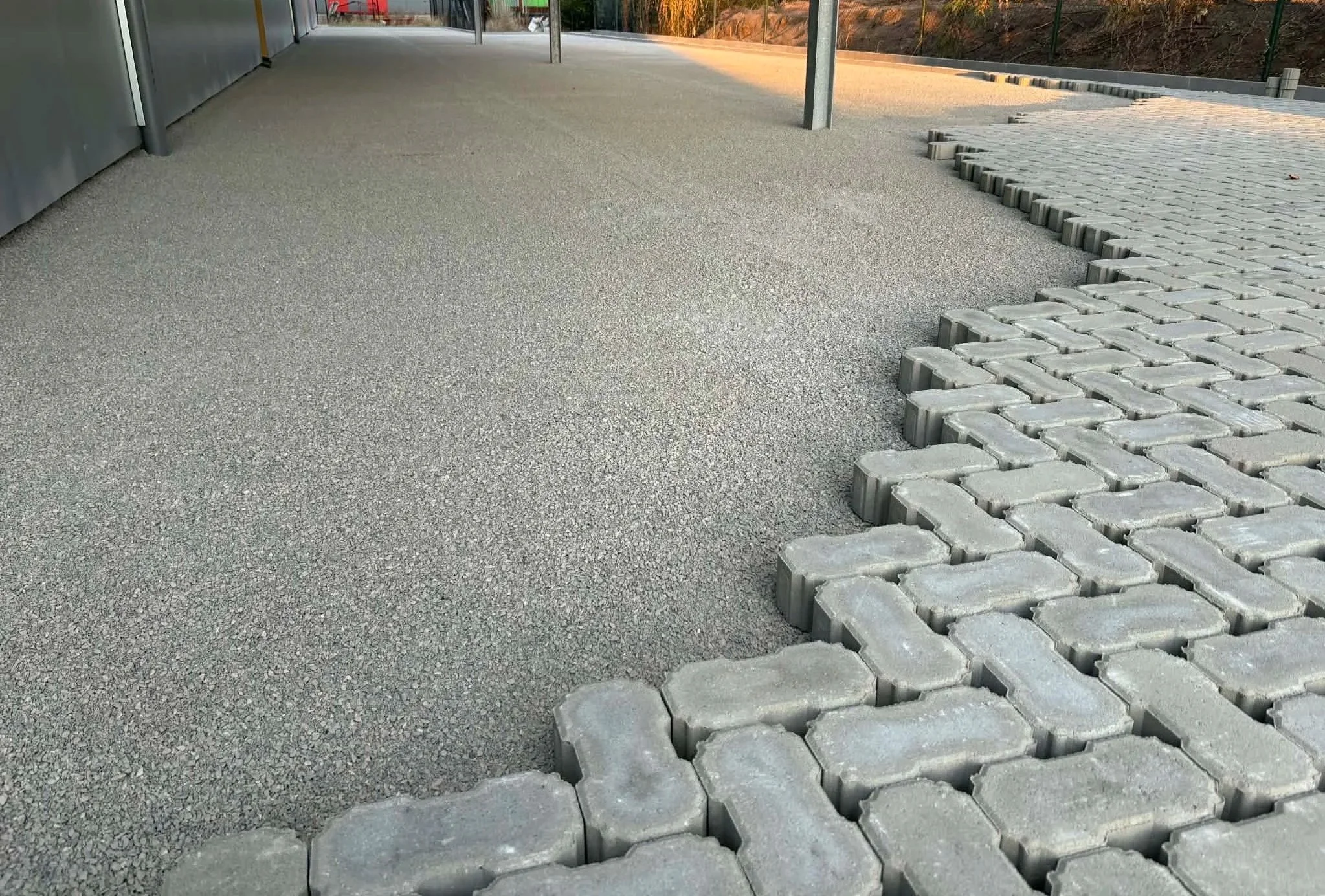De afbeelding toont een betegeld voetpad met een overgang van grindtegels naar betonstenen, ondersteund door metalen palen, onder een afdak.