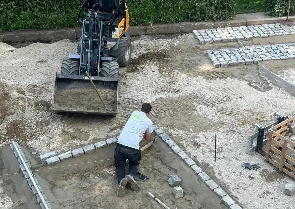 Een persoon die een stenen rand aan een gepland pad legt, terwijl een graafmachine op de achtergrond wordt gebruikt voor grondaankoop en bestratingswerkzaamheden.
project: bestratingwerkzaamheden
locatie: Tessenderlo, Diest, Engsbergen