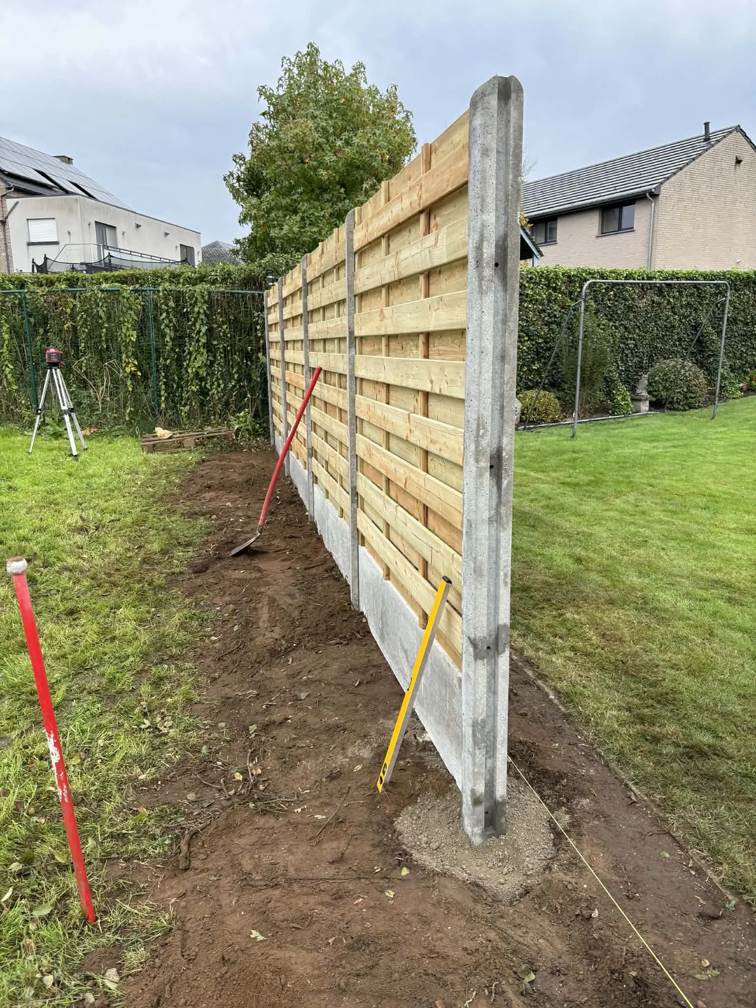 project: beschutting plaatsen, locatie: lammen Twee houten en betonnen afscheidingswanden worden geïnstalleerd in een tuin, met meetlint en gereedschap rondom.