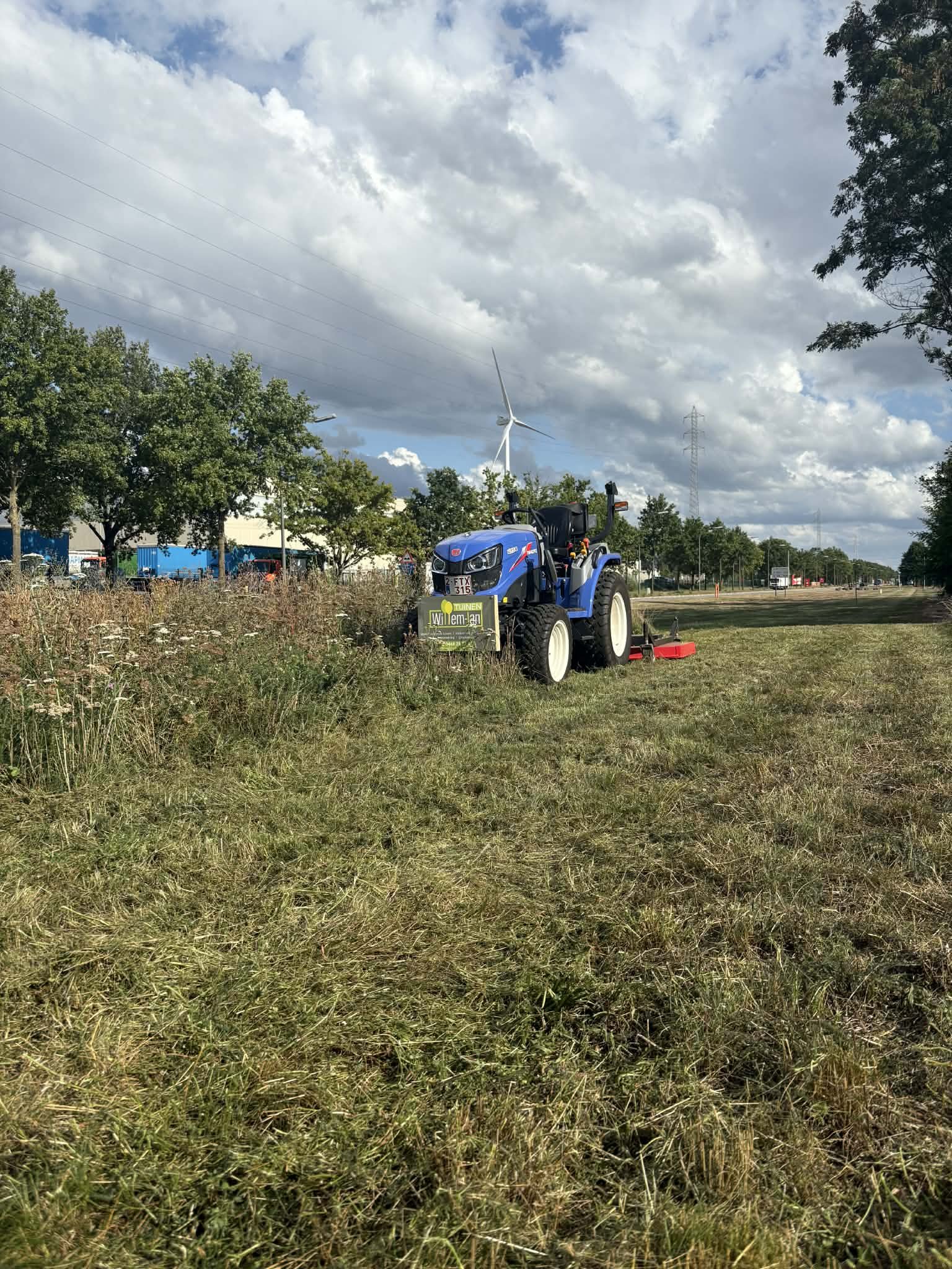 Een veld met gras en bloemen, een blauw tractor met een maaimachine bevindt zich vooraan, er zijn bomen aan de zijkanten en een wolkenlucht boven de horizon.
project: weiland klepelmaaien, tuinaanlegger
locatie: Tessenderlo, Diest, Engsbergen